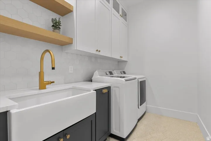 Functional laundry area featuring a white farmhouse sink with a gold-finish faucet, dark base cabinetry, white upper cabinetry, and open wood shelving