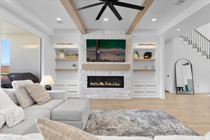 Living area featuring a stone-clad fireplace with a built-in linear firebox, custom built-in cabinetry with floating wood shelves, wood-finish flooring, exposed ceiling beams, and a modern ceiling fan