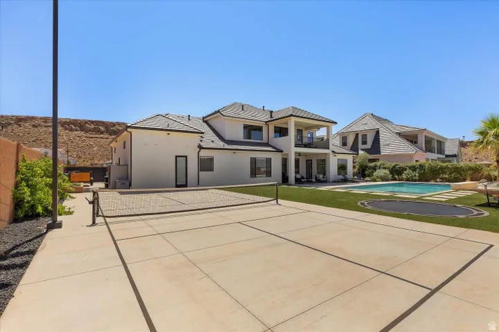 Expansive concrete sports court featuring a net, alongside a manicured lawn with an in-ground swimming pool and an integrated trampoline