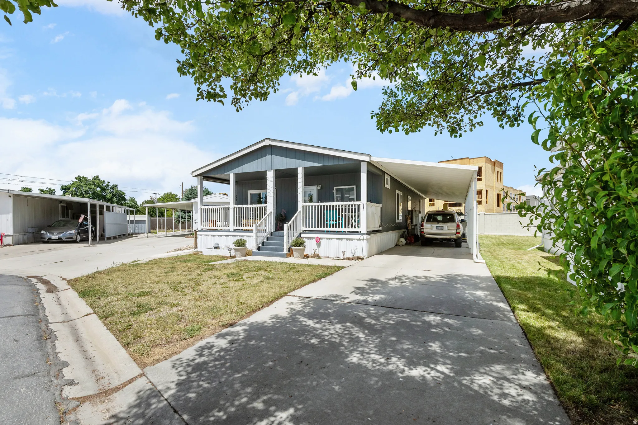 Property exterior featuring a covered front porch with white railing, gray siding, and a paved driveway leading to a car port