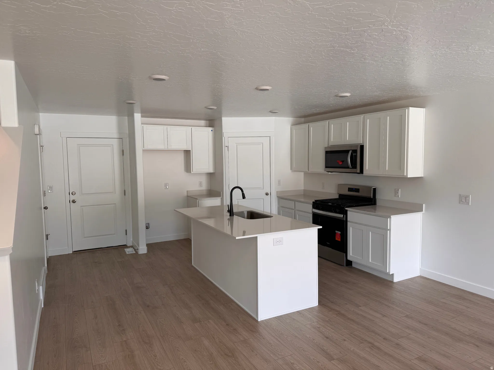 Kitchen featuring wood-finish flooring, white shaker cabinetry, light-toned countertops, a stainless steel gas range, and a central island with a sink and matte black faucet