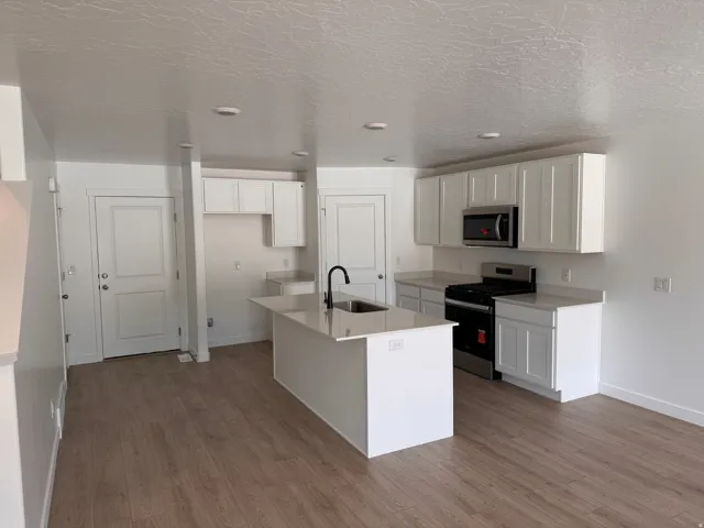 Kitchen featuring wood-finish flooring, white shaker cabinetry, light-toned countertops, a stainless steel gas range, and a central island with a sink and matte black faucet