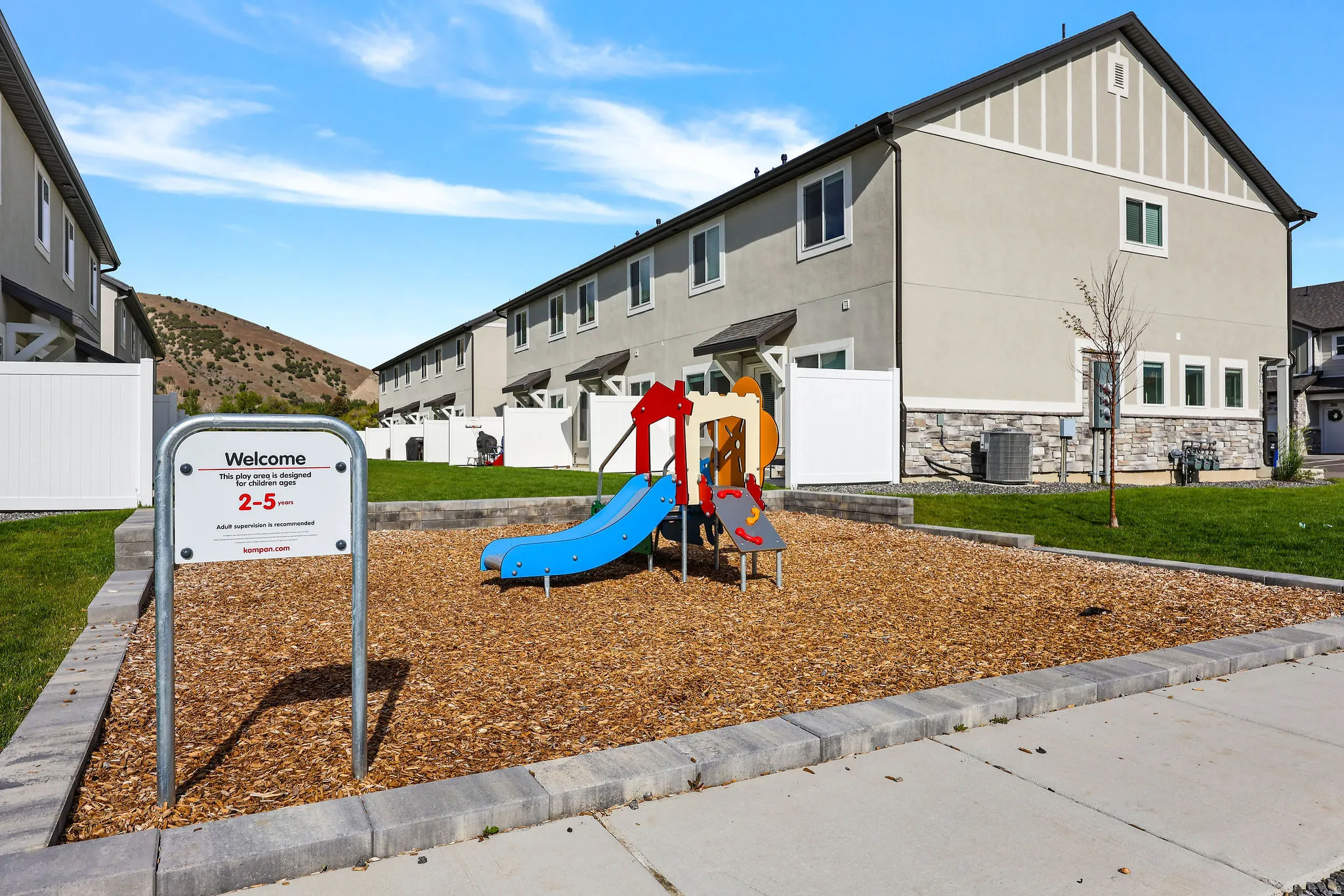 Community playground featuring a colorful playset on wood chip surfacing, bordered by a concrete block retaining wall