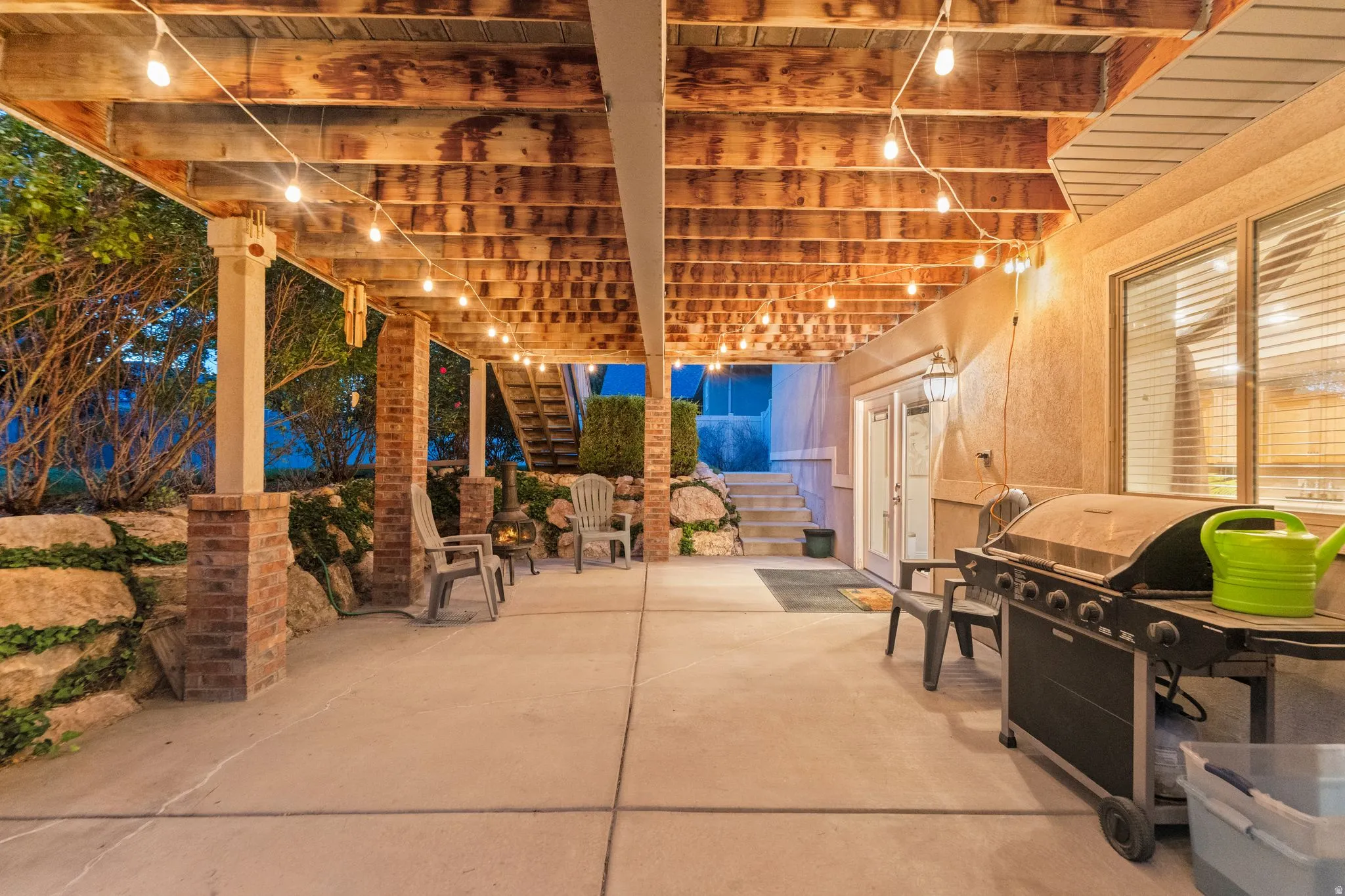 Covered concrete patio featuring string lighting, brick column accents, and stone retaining walls