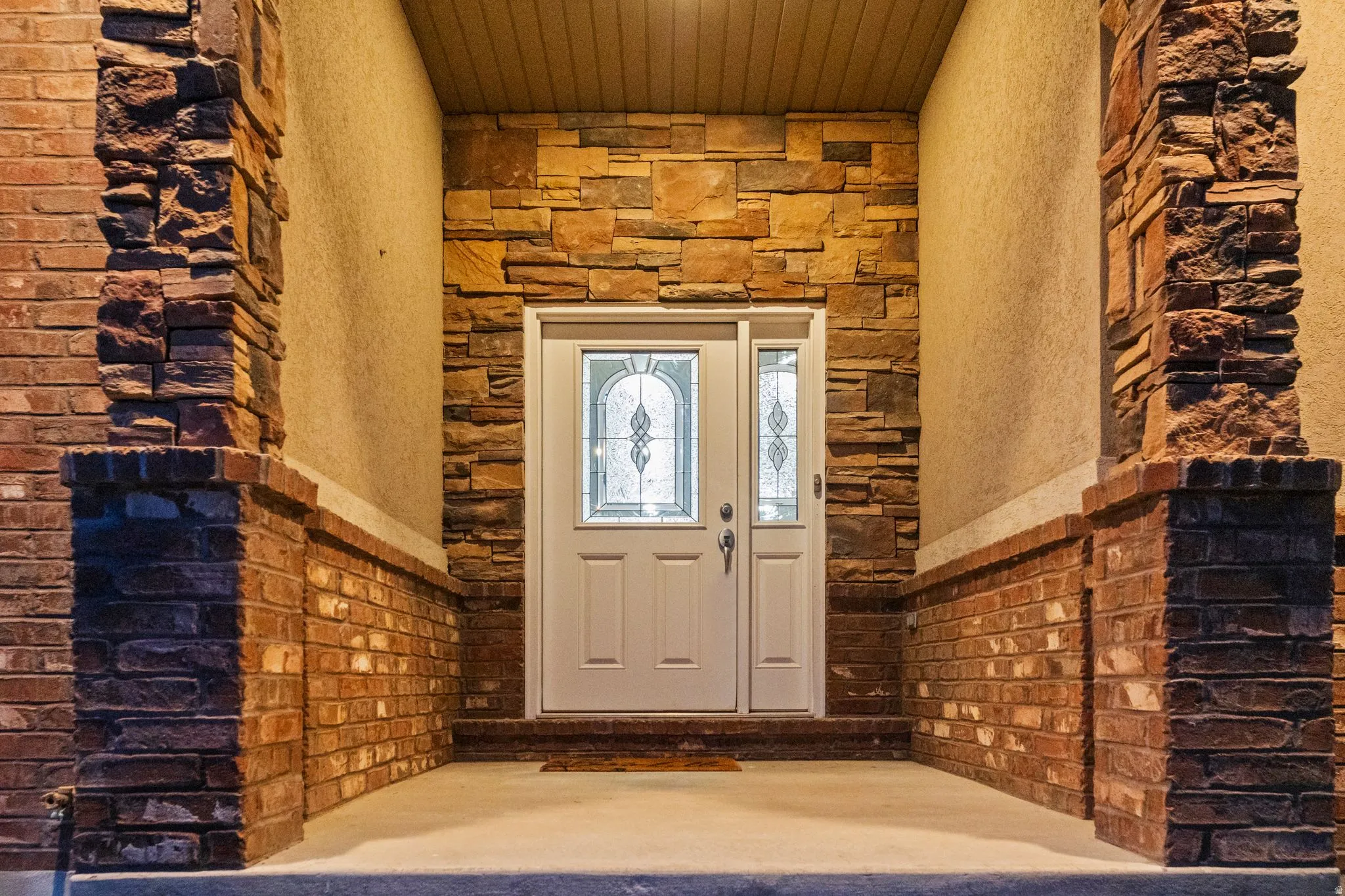 Entryway featuring a white door with decorative glass, stone veneer, brick detailing, and a stucco finish