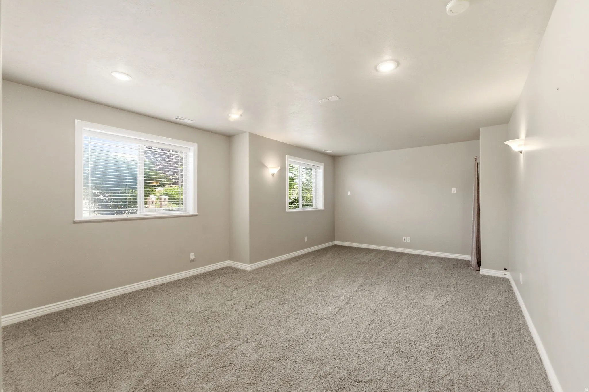 Spacious room featuring neutral carpeting, recessed ceiling lighting, two windows with blinds, and light-toned wall sconces