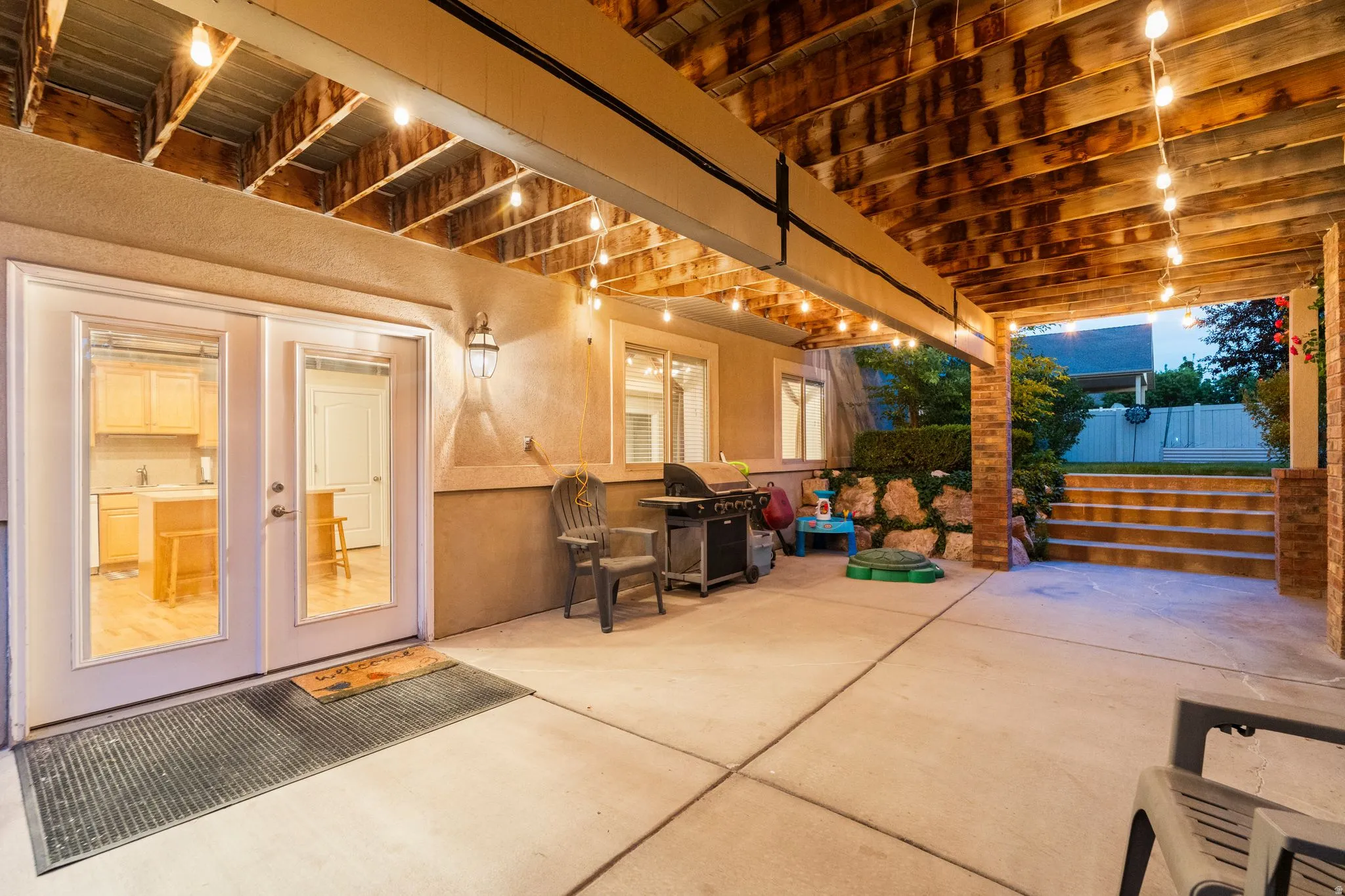 Covered concrete patio with string lighting, brick pillars, and built-in steps leading to the upper yard