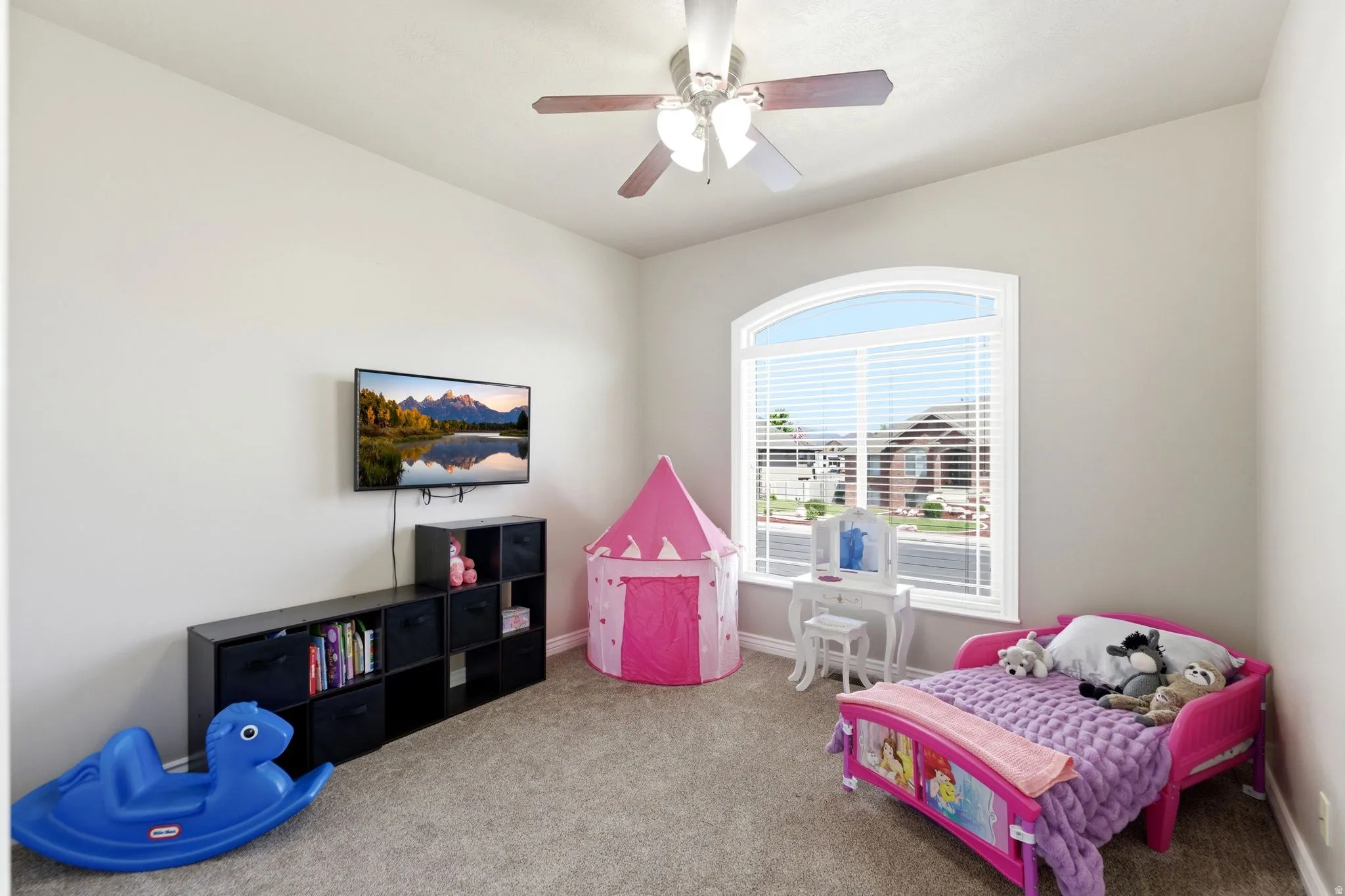 Room featuring light gray wall paint, neutral carpeting, an arched window with blinds, a ceiling fan with light fixture, and a wall-mounted television