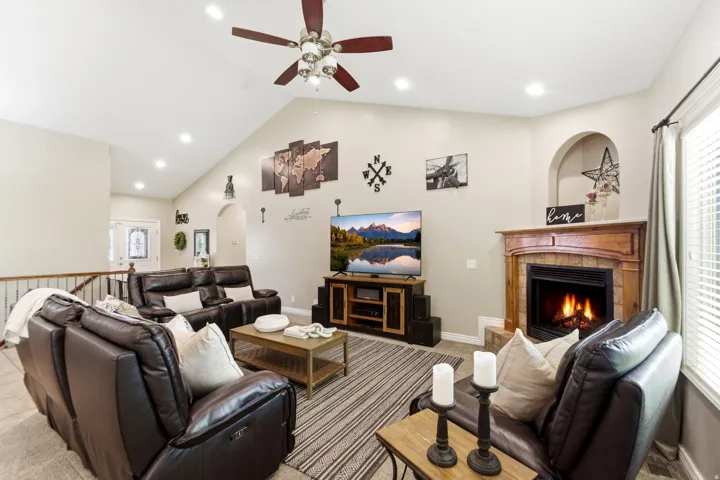 Spacious living area featuring a vaulted ceiling with recessed lighting and a ceiling fan, a fireplace with a wood mantel and stone surround, and large windows with blinds
