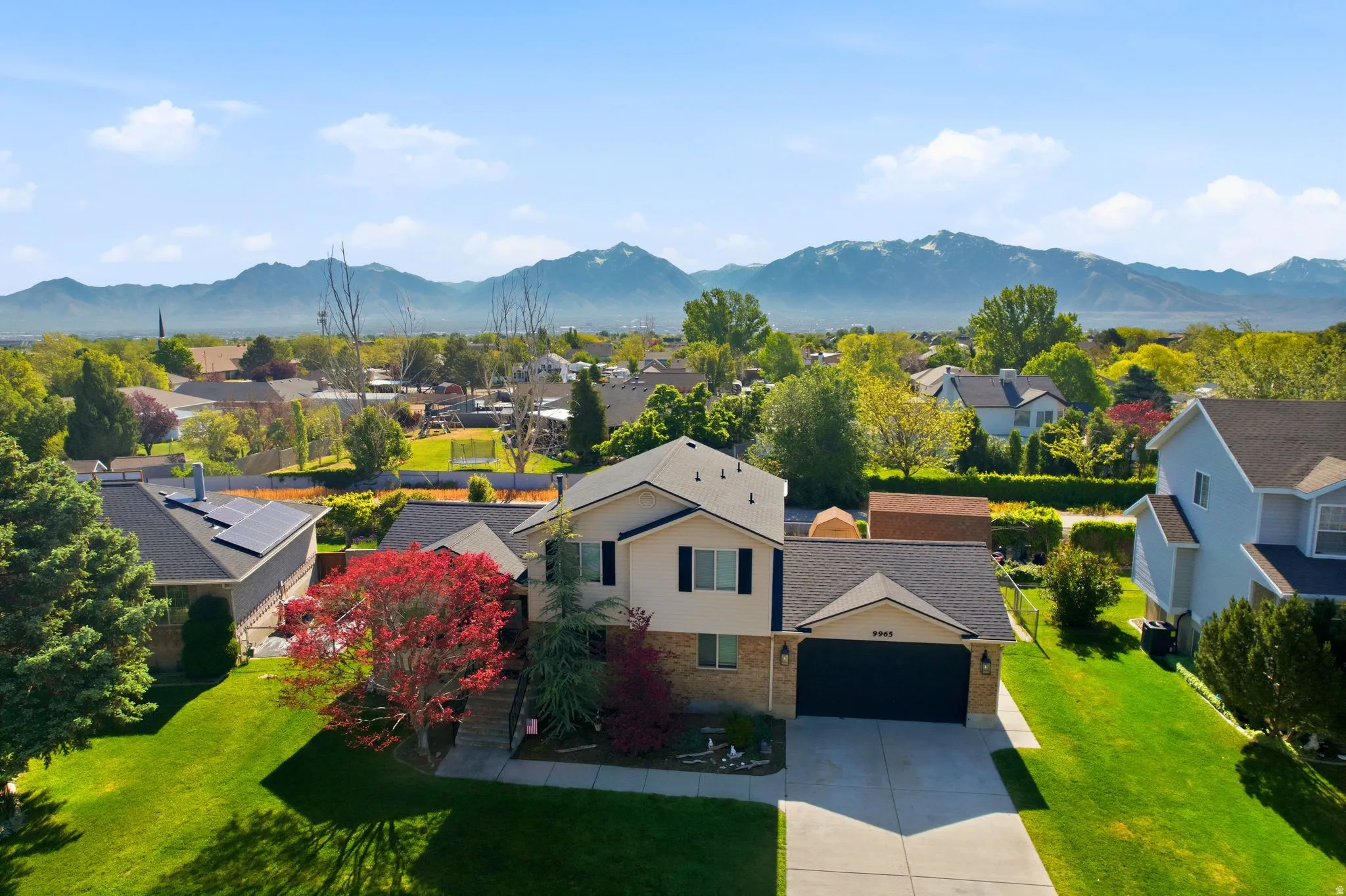 Two-story residence featuring a two-car garage, manicured front lawn, and mature trees, set against a distant mountain backdrop