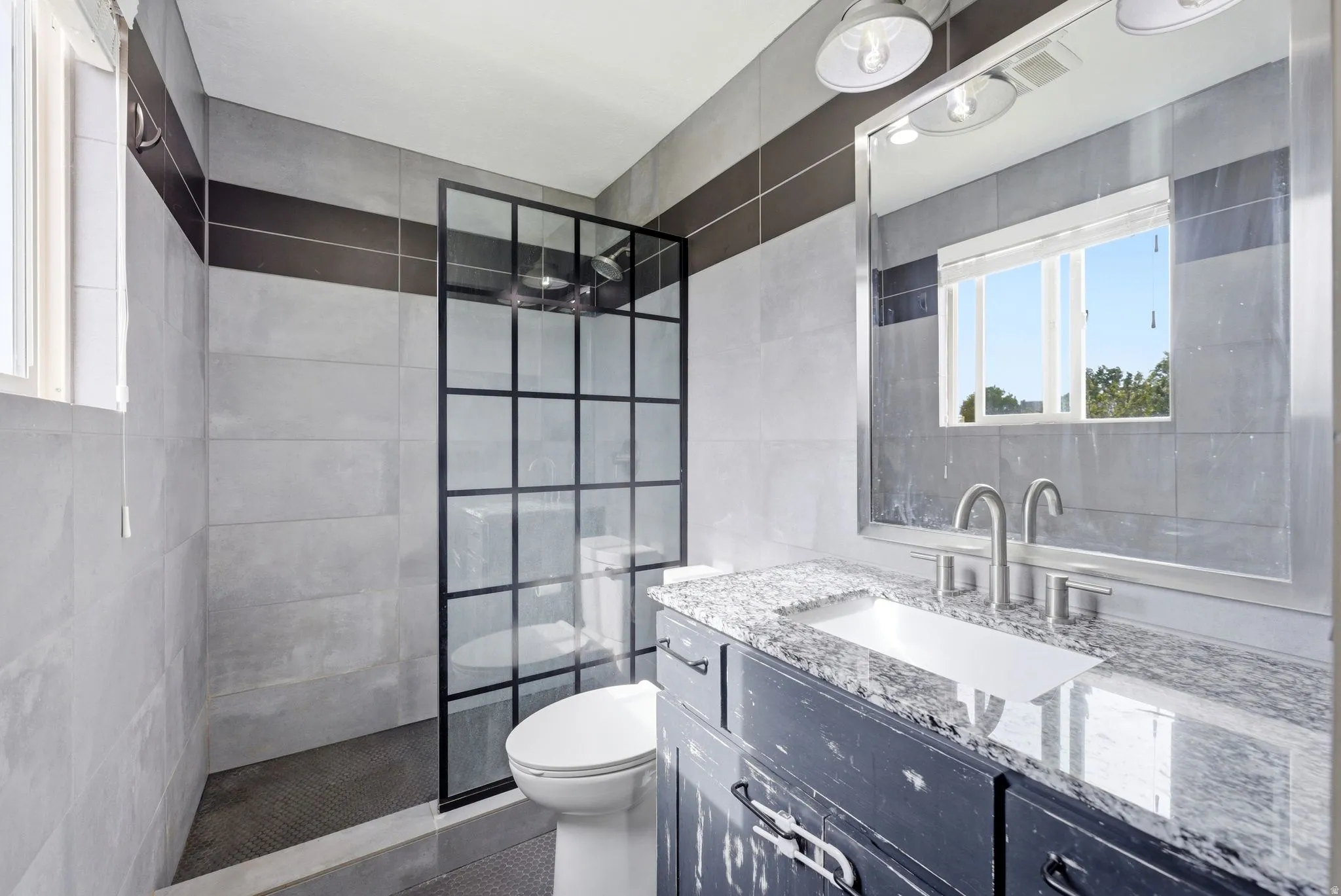 Bathroom featuring a grid-pattern glass shower enclosure, dark grey vanity with a light-toned countertop, rectangular undermount sink, and brushed nickel fixtures