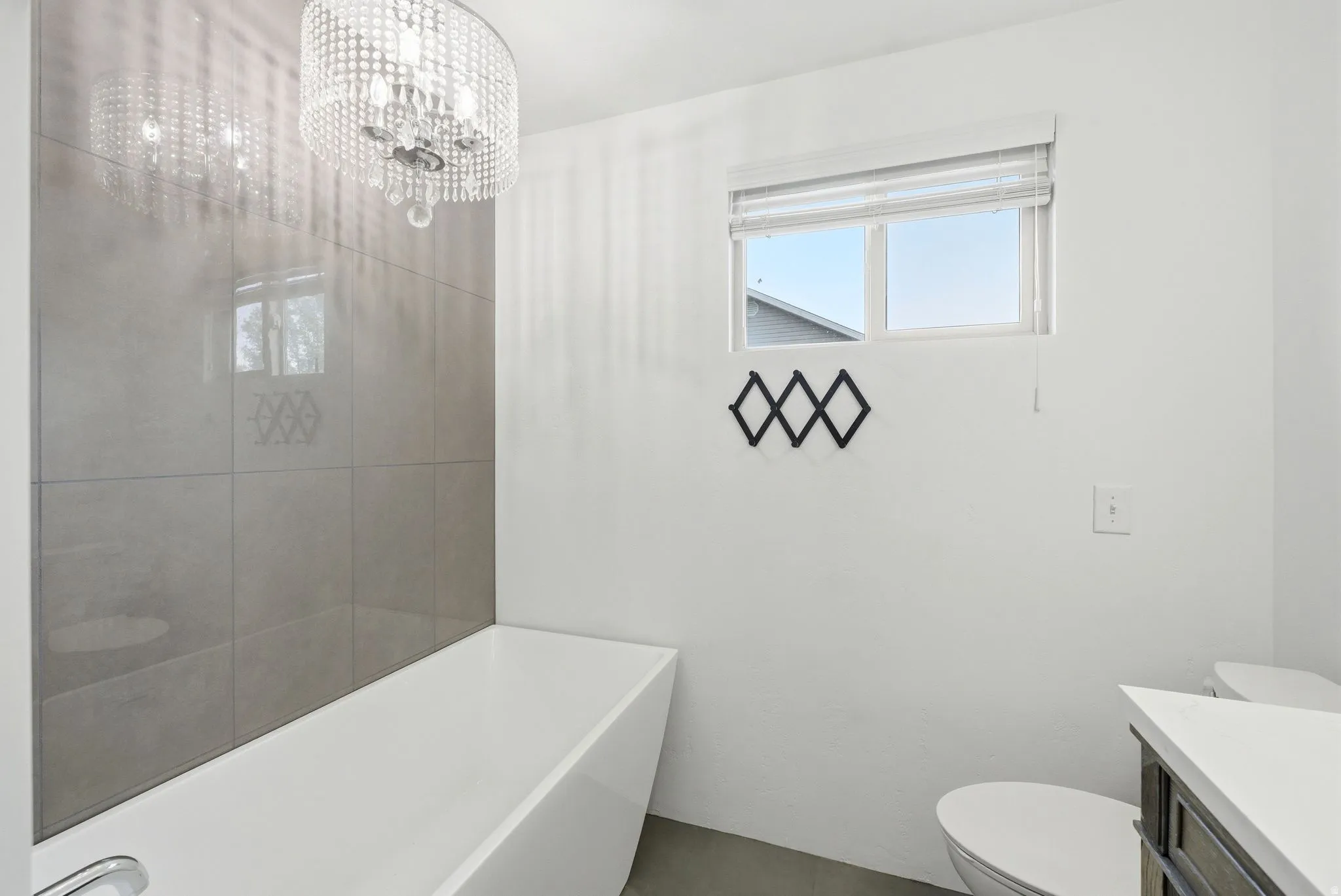 Freestanding rectangular bathtub with a chrome faucet, large format grey wall tiling, and a crystal chandelier