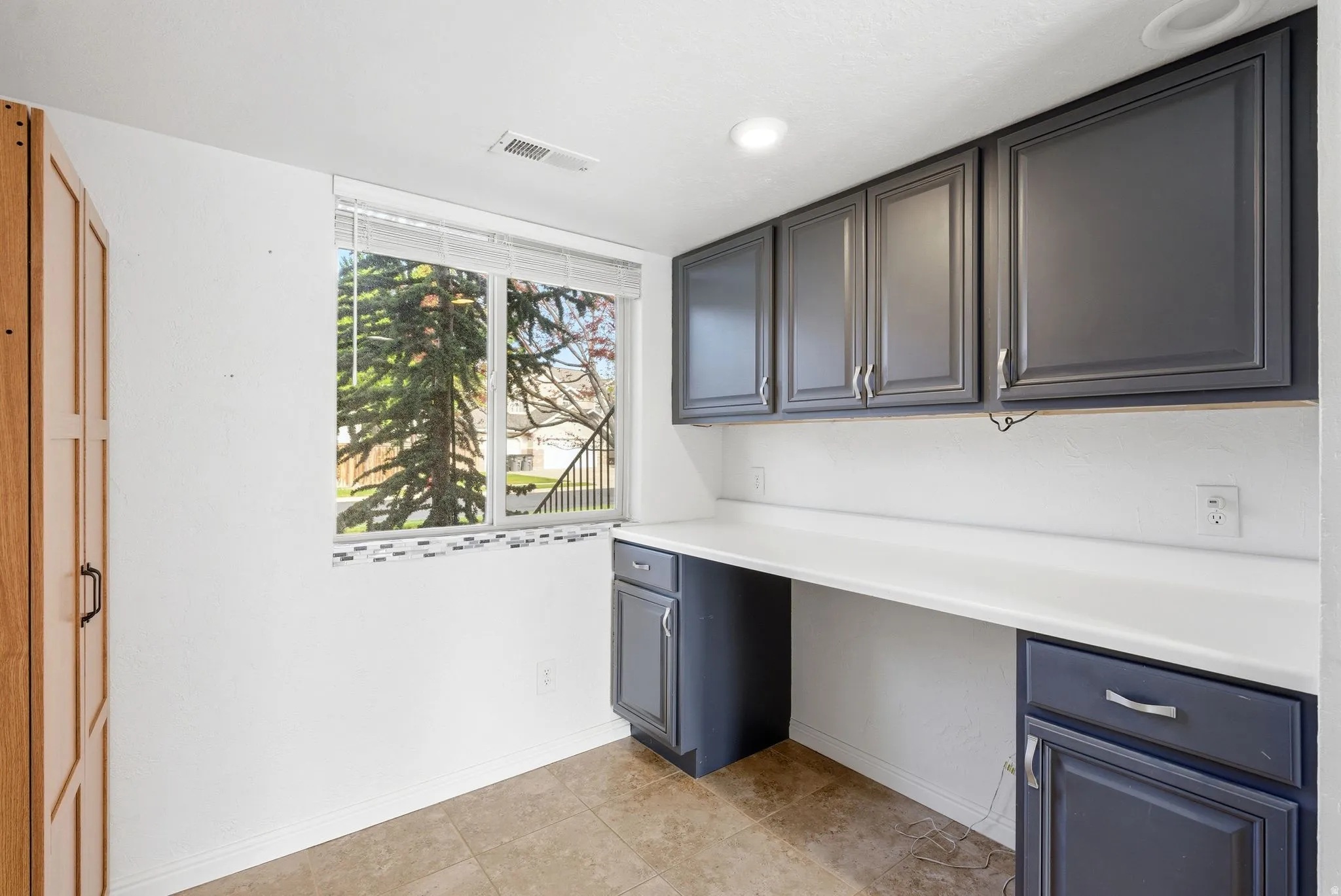 Functional utility area featuring a window, tile flooring, built-in desk with a white countertop, and dark cabinetry