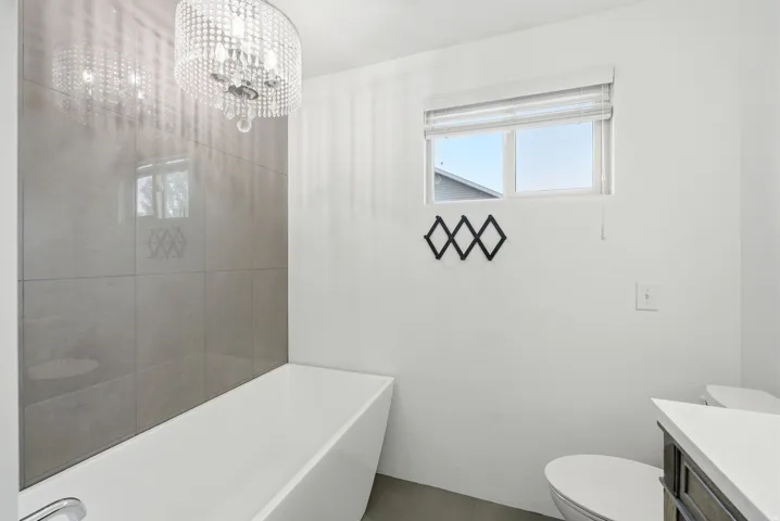 Freestanding rectangular bathtub with a chrome faucet, large format grey wall tiling, and a crystal chandelier