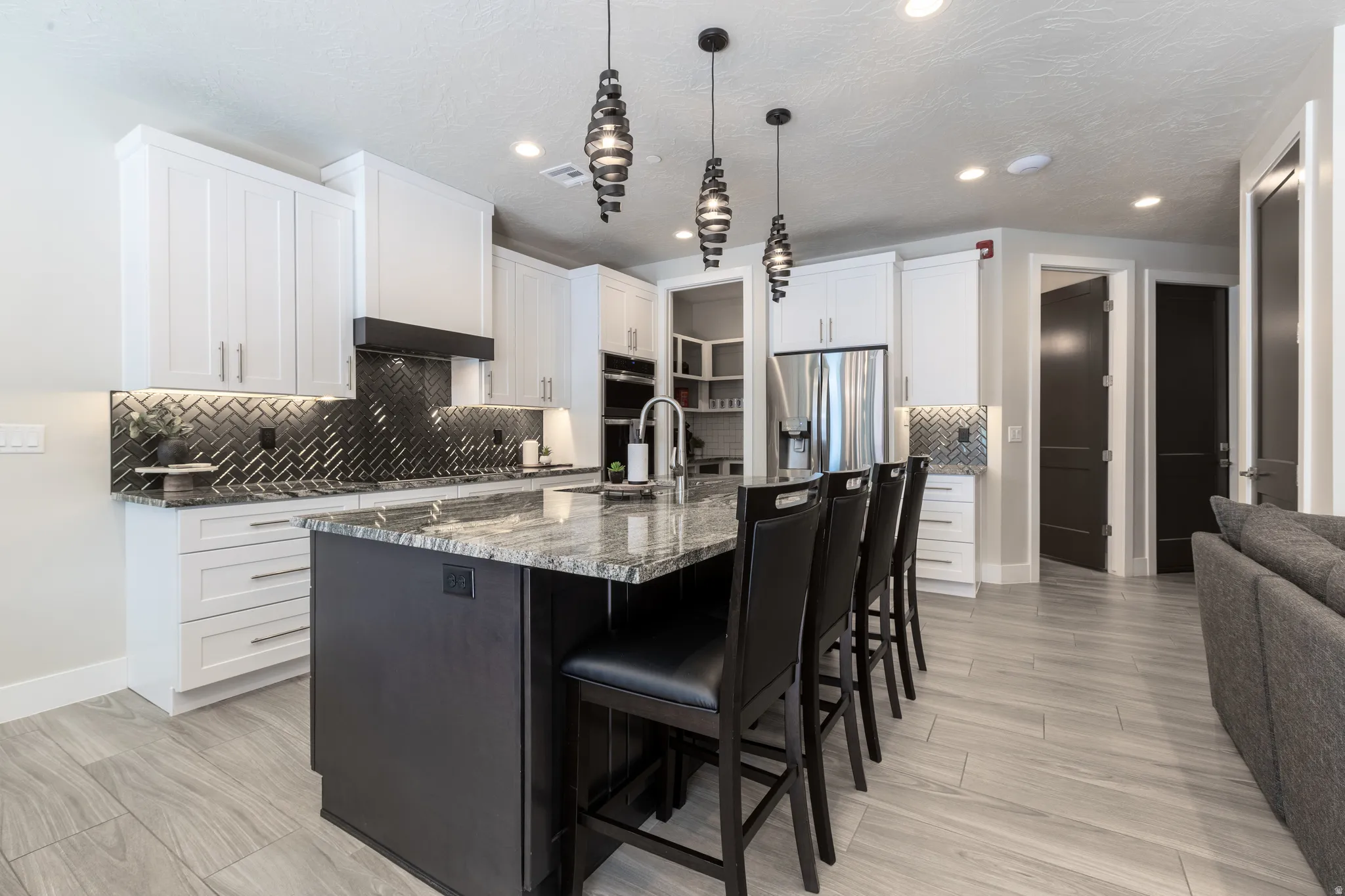 Gourmet kitchen featuring a large central island with seating, granite countertops, white cabinetry, dark herringbone tile backsplash, and stainless steel appliances
