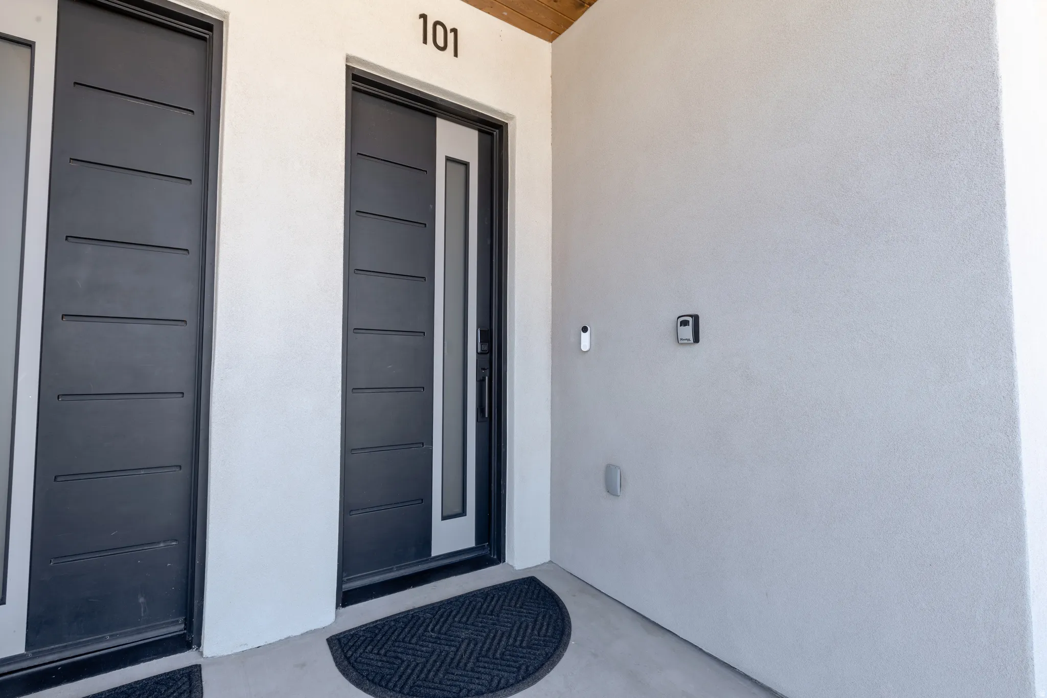 Modern entrance featuring a dark gray door with horizontal paneling and a vertical frosted glass insert, complemented by a white stucco exterior and black house numbers