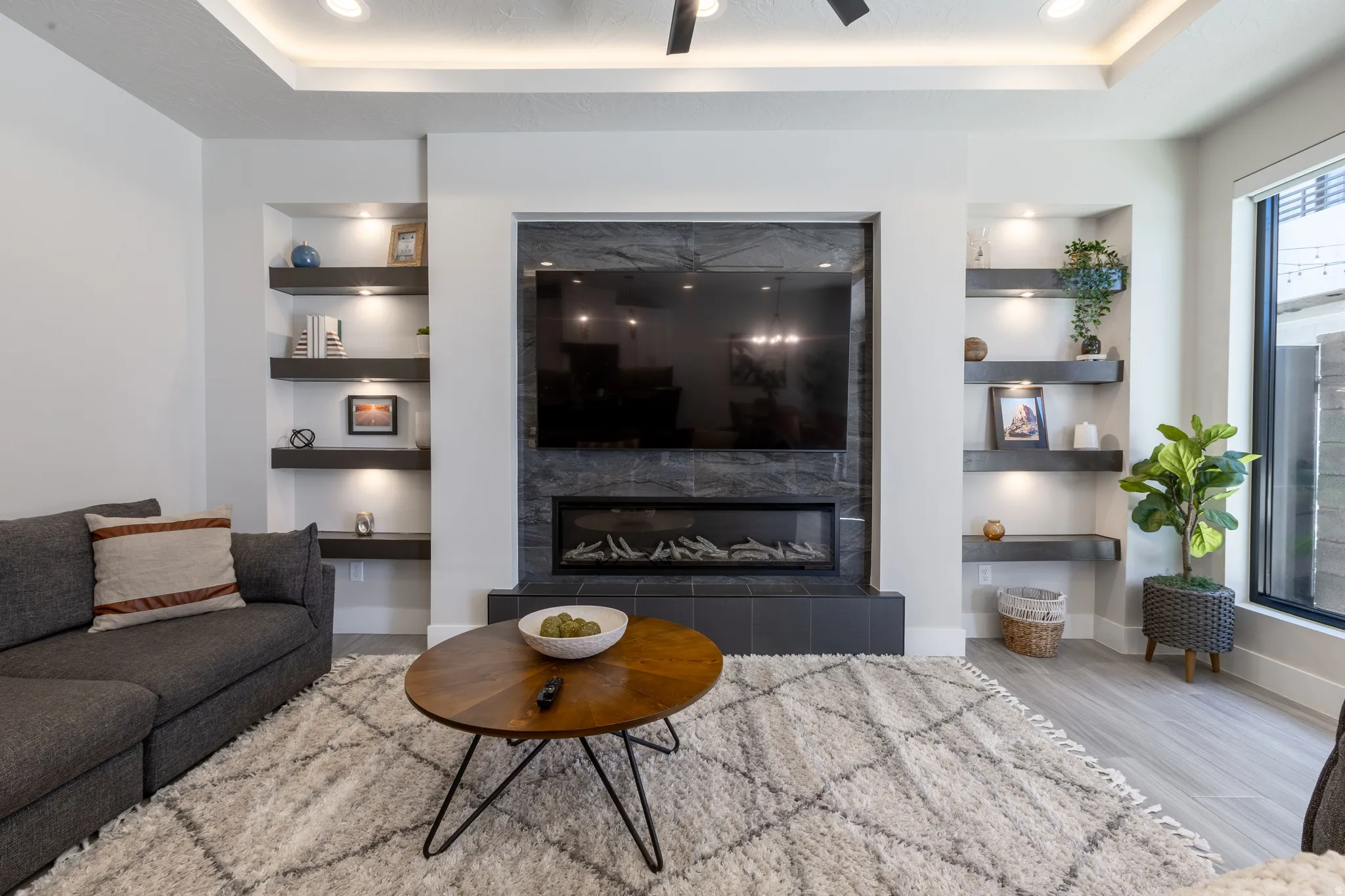Contemporary living area featuring a linear fireplace with a dark stone-look surround, integrated display niches with accent lighting, a tray ceiling with recessed lighting, and light wood-finish flooring