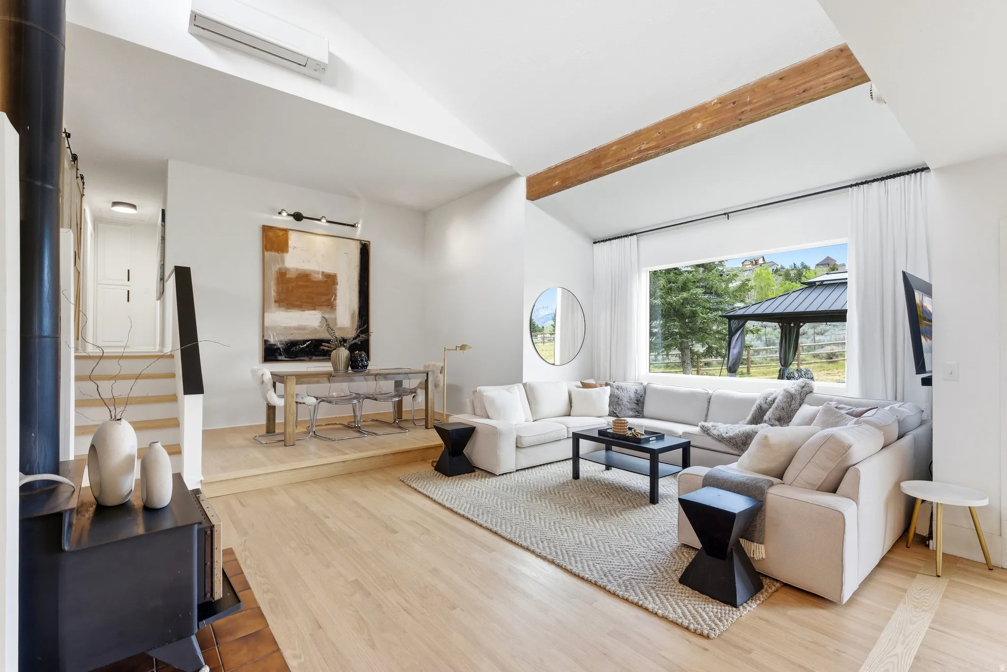 Spacious living area featuring light wood-finish flooring, a prominent wooden beam, and a large window overlooking a gazebo and natural landscape