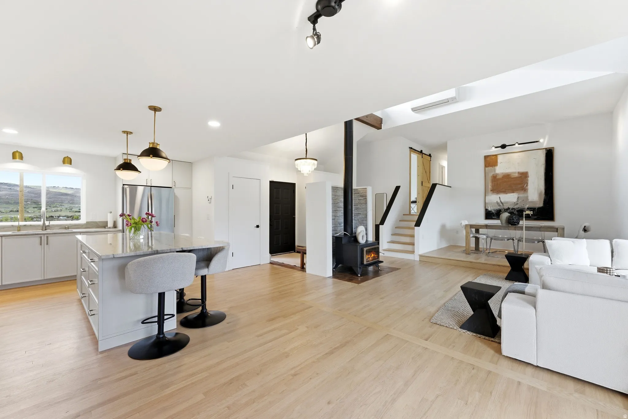 Open-concept living area featuring light wood-finish flooring, a modern kitchen island with pendant lighting, a wood-burning stove, and a staircase with a black handrail