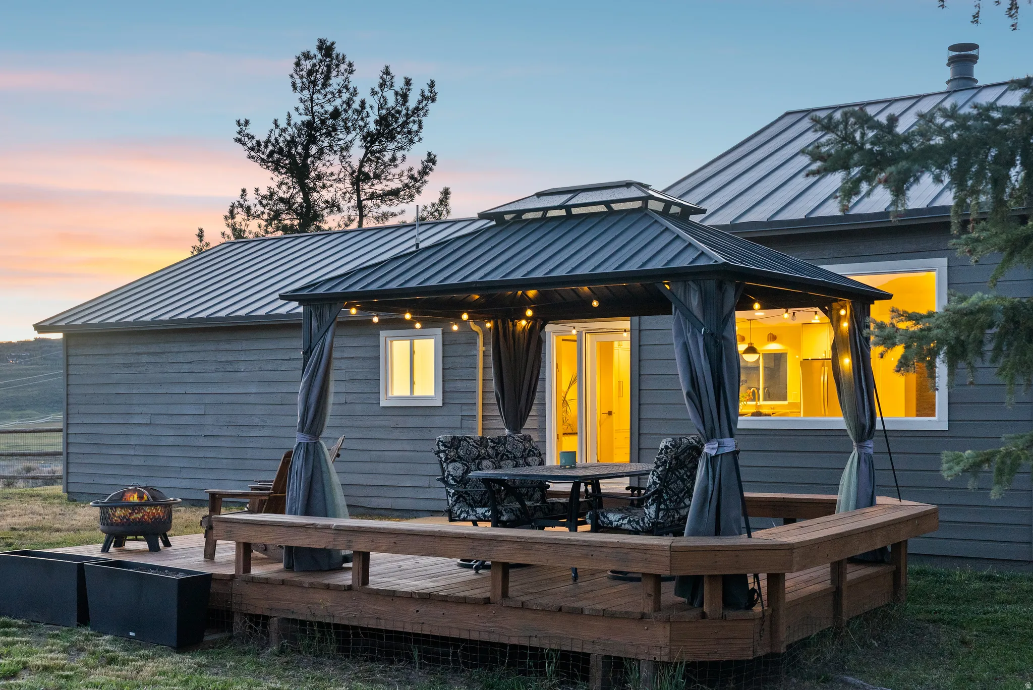 Expansive wood deck featuring a covered gazebo with string lights, integrated seating, and a nearby fire pit