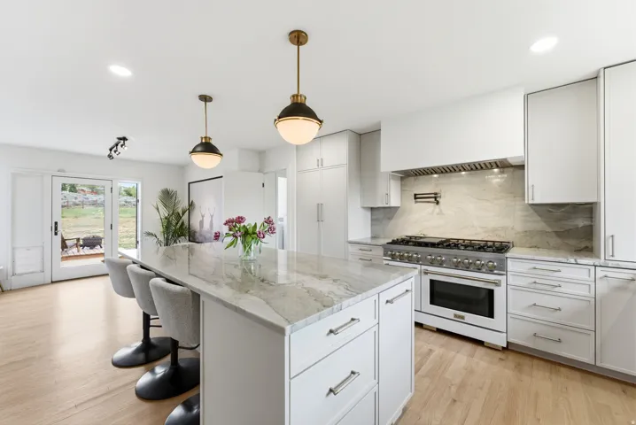 Bright kitchen featuring a substantial island with a stone countertop, a professional-grade range, a complementary stone backsplash, white cabinetry, and wood-finish flooring