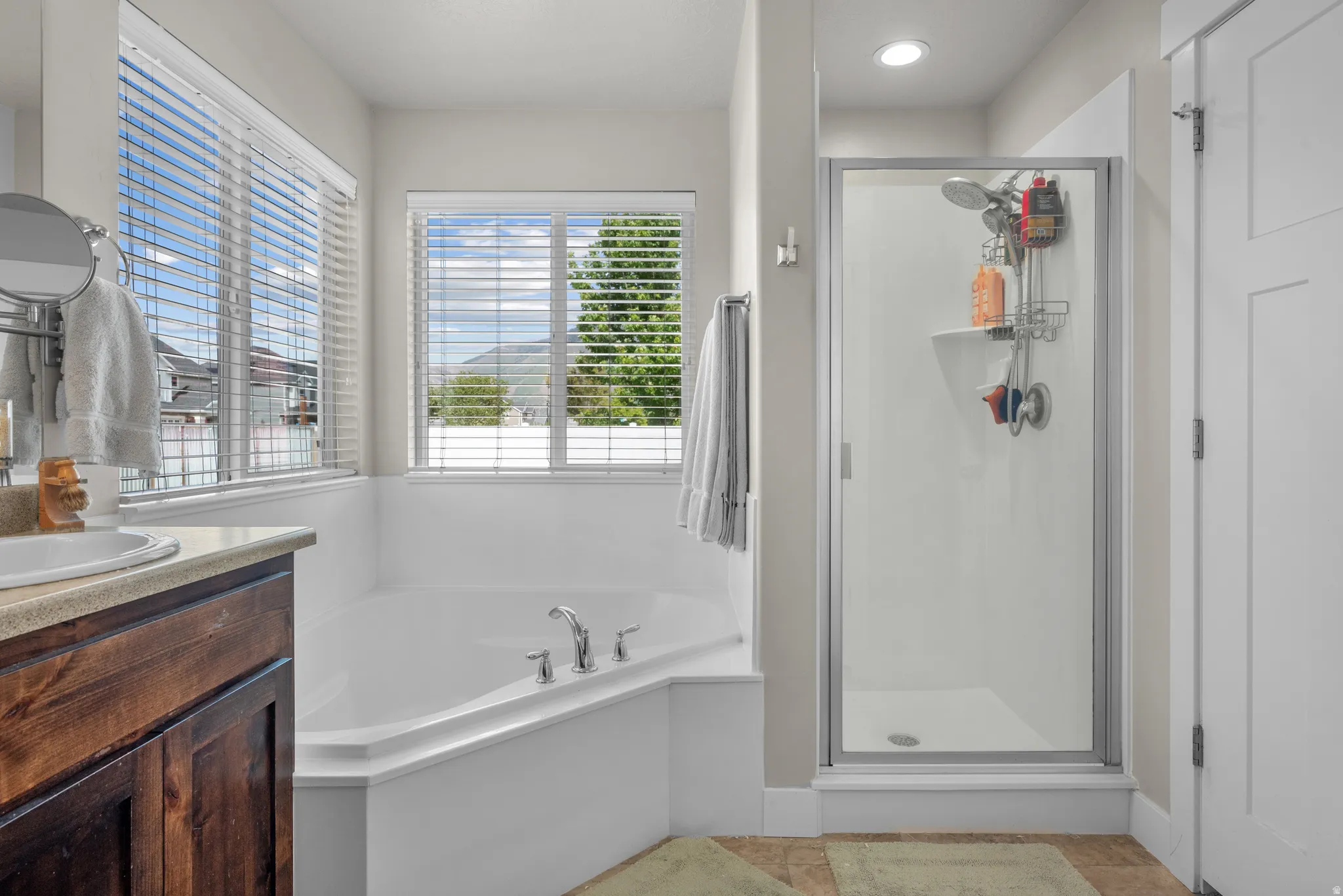 Soaking tub with chrome fixtures, glass-enclosed shower, wood-finish vanity with countertop, and double windows with blinds