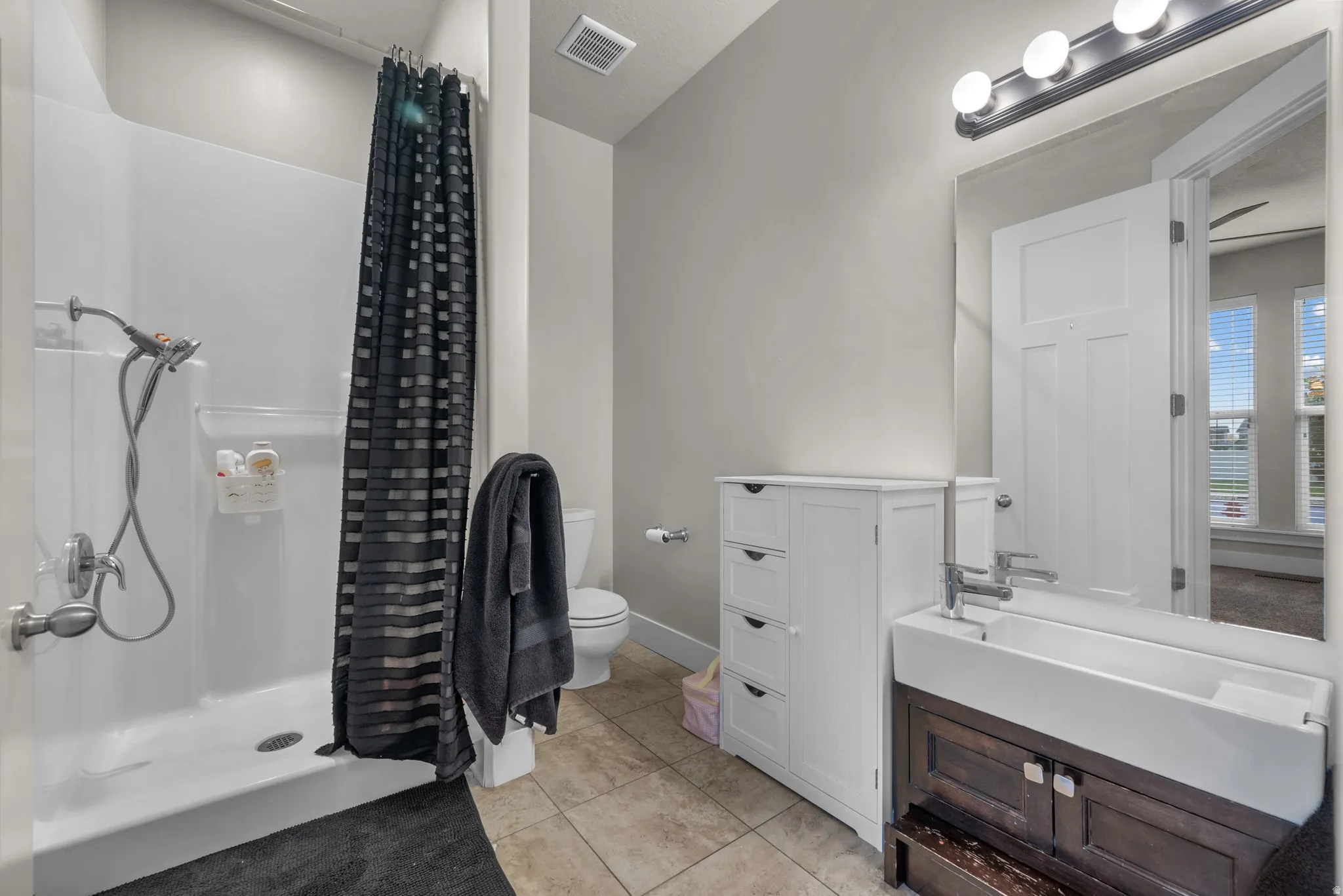Contemporary bathroom featuring a white single-piece shower surround, a dark wood-finish vanity with a vessel sink, a chrome faucet, a large frameless mirror, and neutral-toned tile flooring