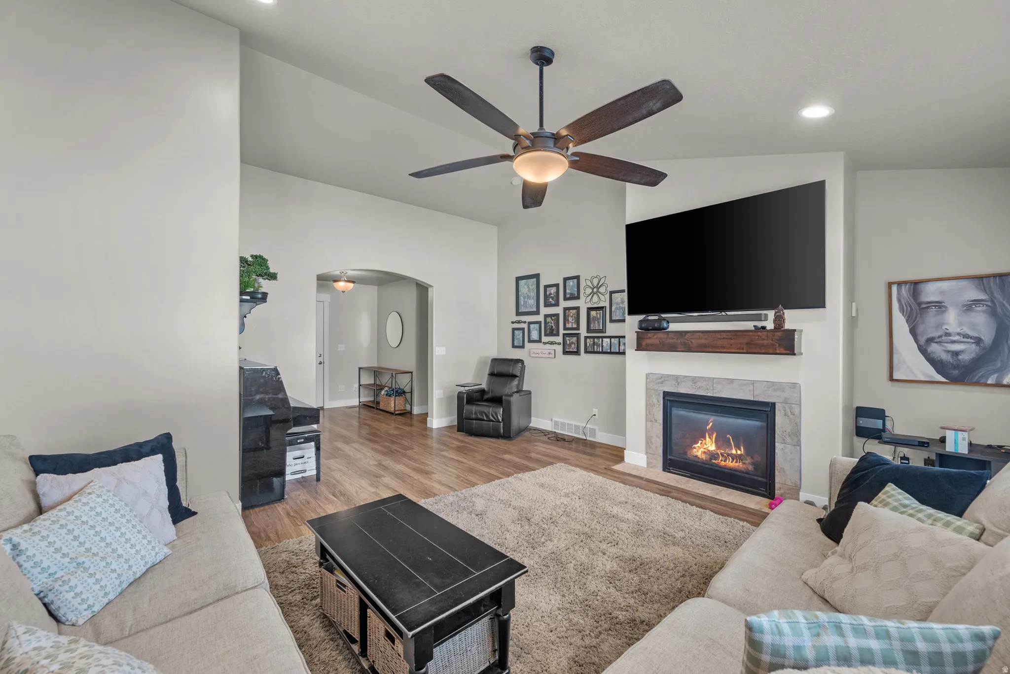 Living area featuring wood-finish flooring, a ceiling fan with integrated lighting, recessed lighting, an arched doorway, and a fireplace with a stone-look surround and wood mantel