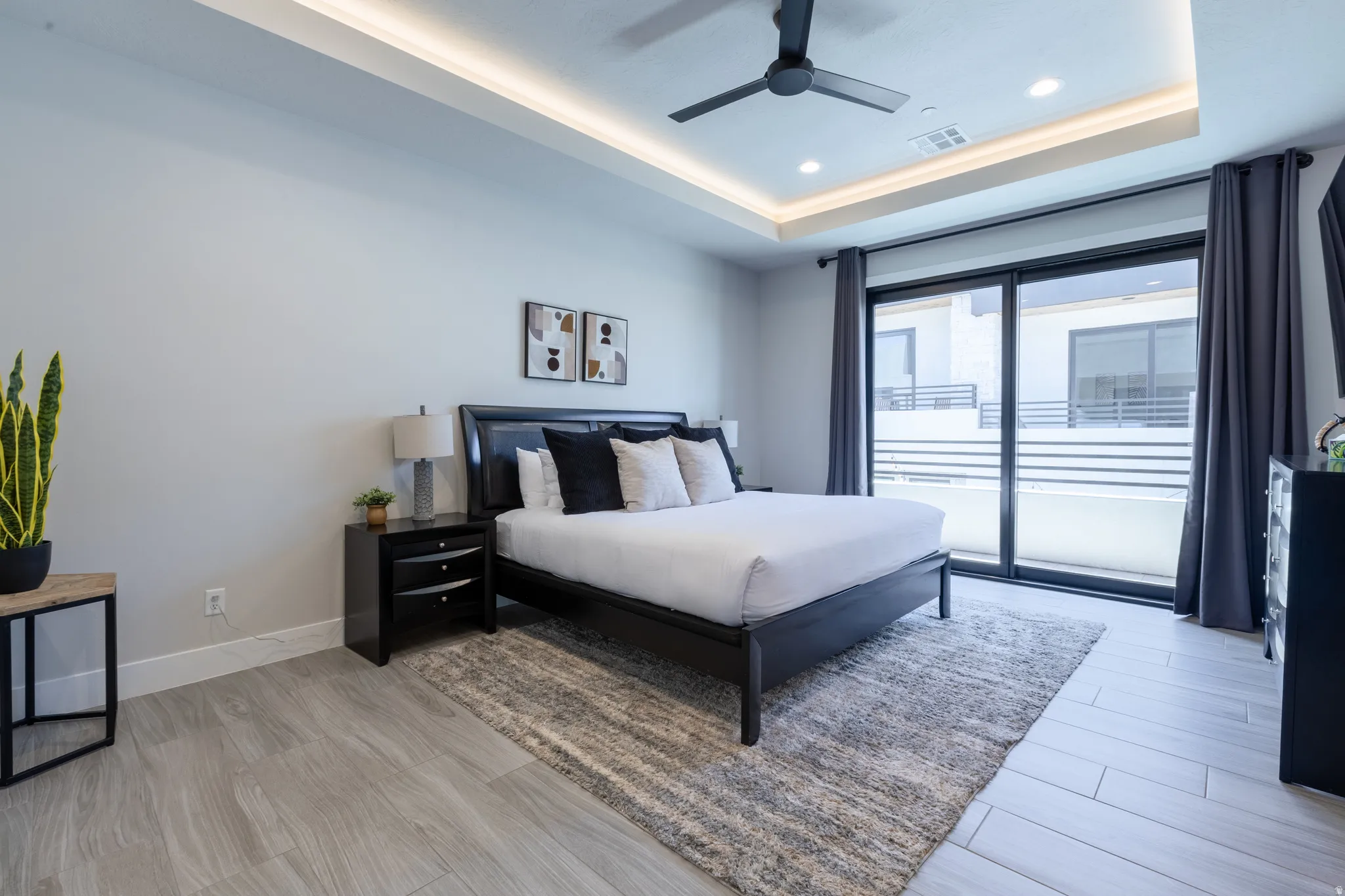 Recessed tray ceiling with integrated lighting, modern ceiling fan, sliding glass door with dark drapery, wood-finish flooring, and black nightstands