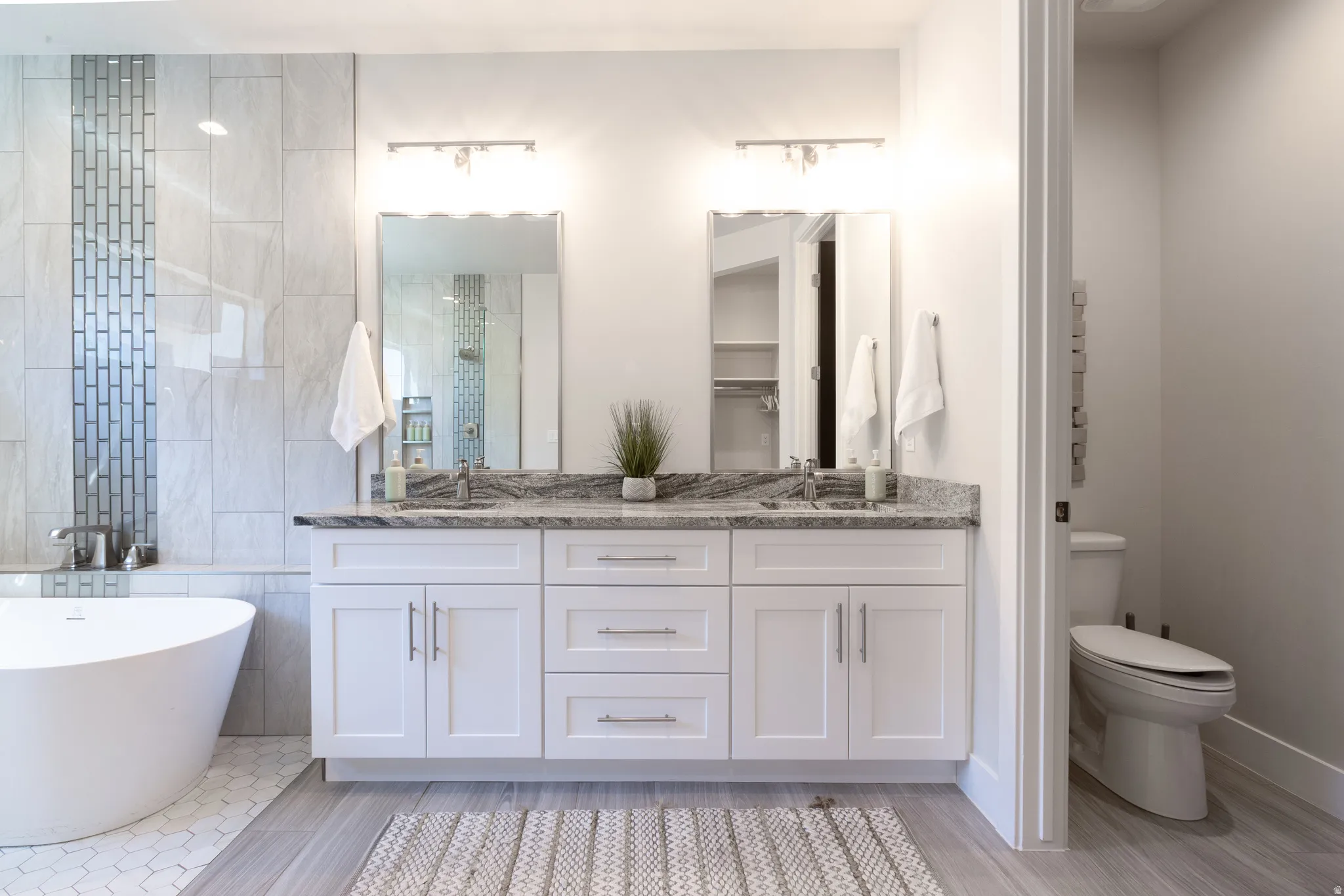 Spacious bathroom featuring a dual vanity with white cabinetry and a granite-style countertop, a freestanding soaking tub with hexagonal tile surround, a large mirror, and wood-finish flooring