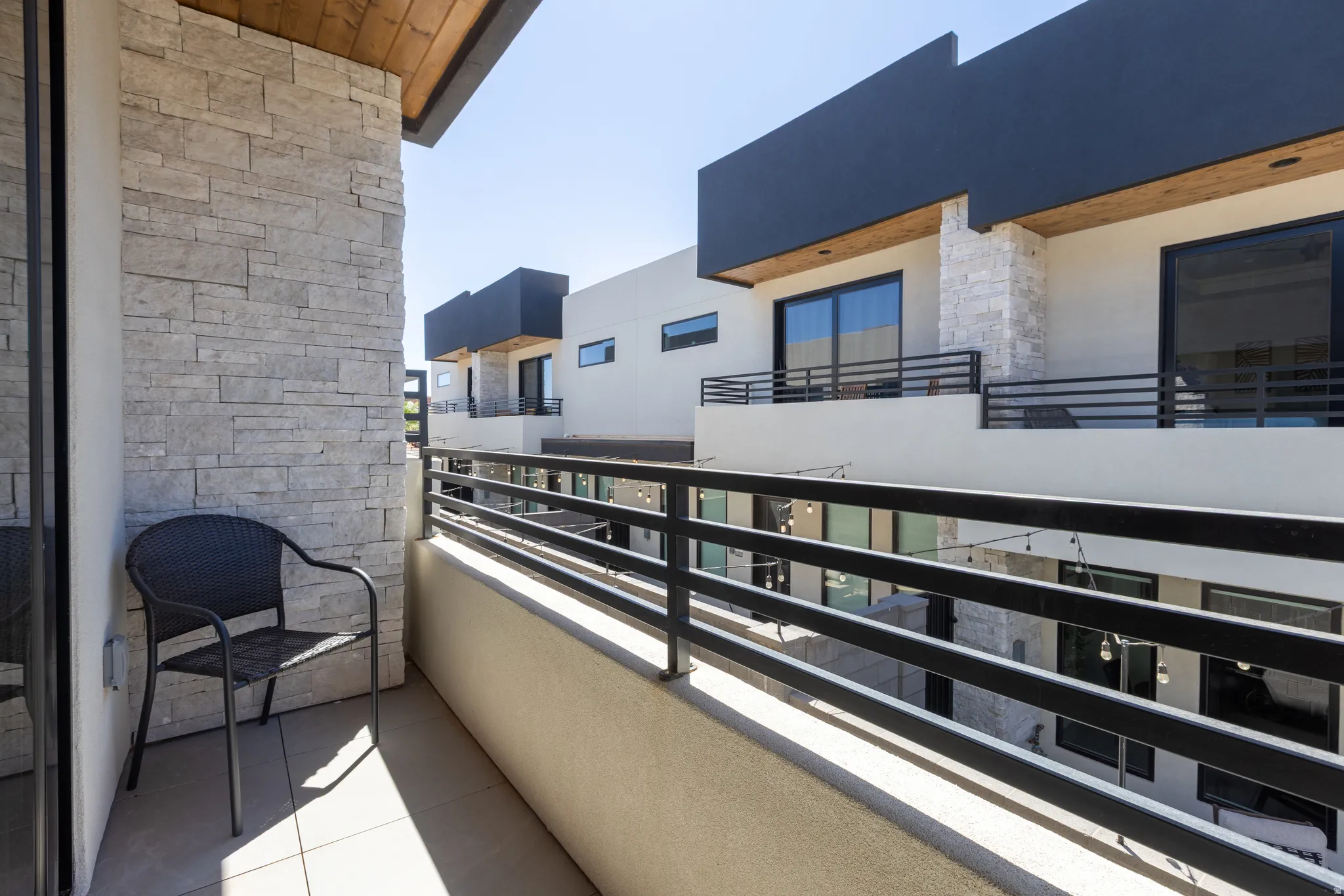 Private balcony with tile flooring, featuring a stacked stone accent wall, black metal railing, and wood-finish ceiling accents