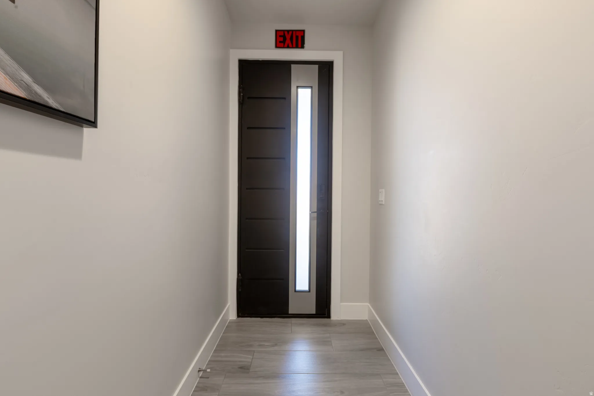 Hallway featuring light gray walls, wood-finish flooring, white baseboards, and a modern dark-paneled door with an integrated vertical frosted glass panel