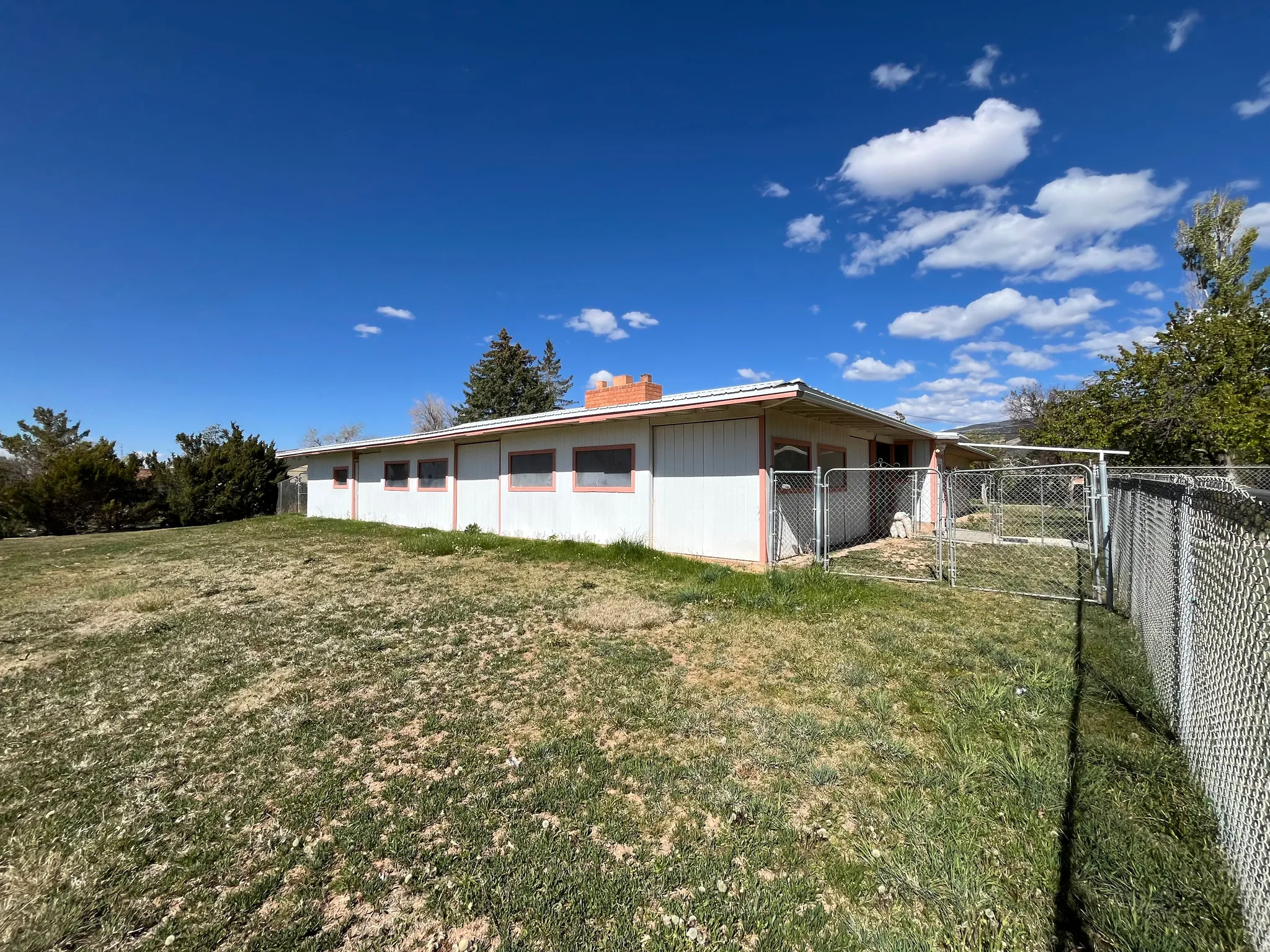 Exterior featuring white siding with pink trim, multiple windows, a chain-link fenced area, and a large grassy yard