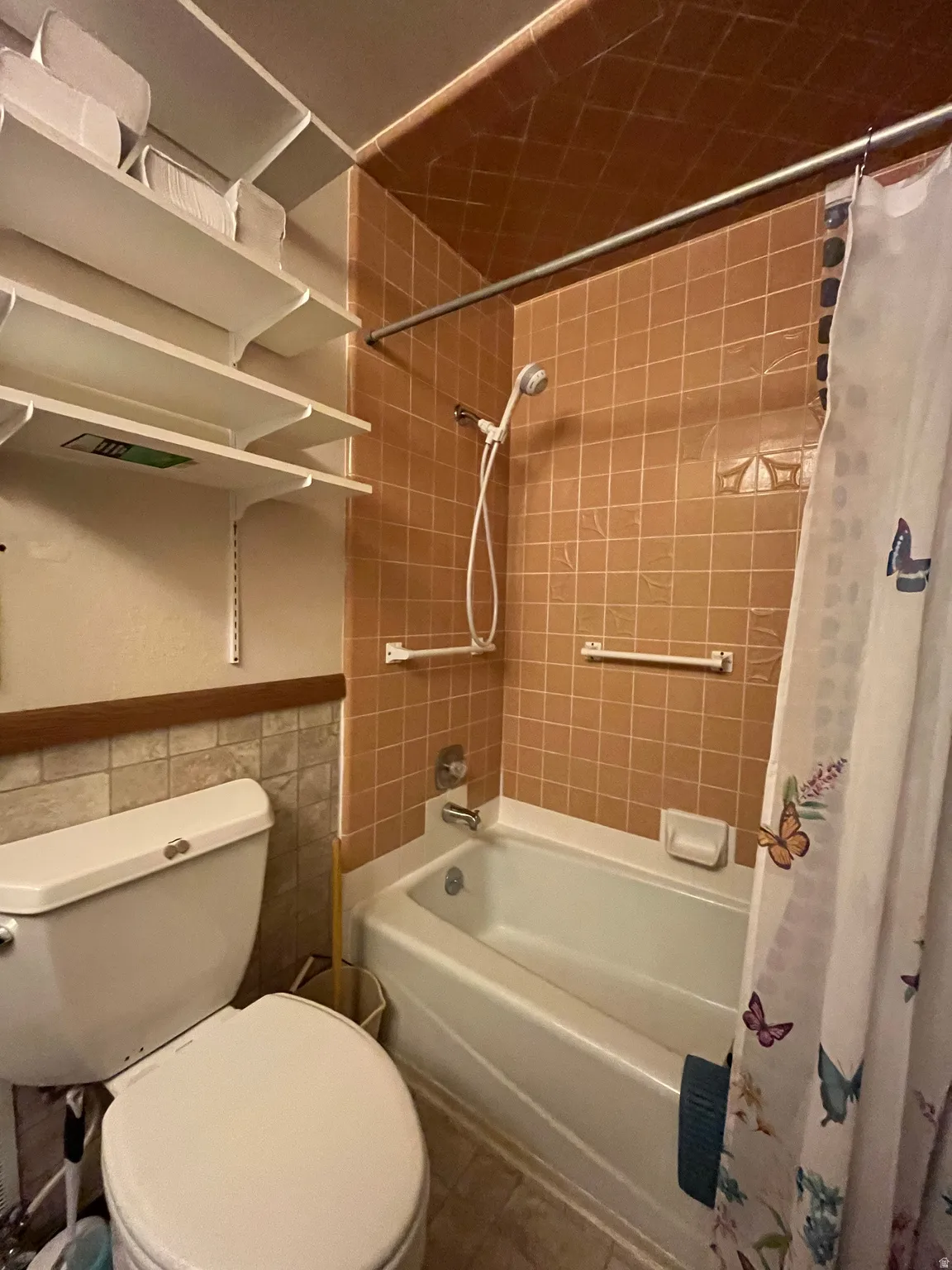 Bathroom featuring a white bathtub, ceramic tile surround, wall-mounted showerhead with handheld attachment, and white shelving units