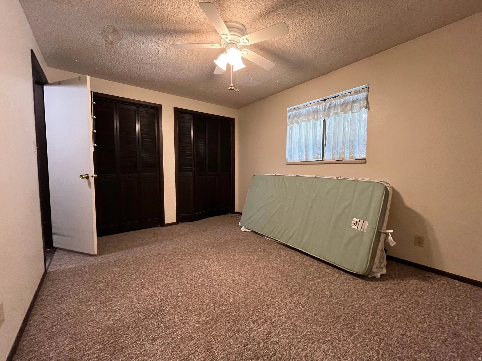 Carpeted room featuring a ceiling fan with light fixture, two louvered bifold closet doors, and a window with curtains