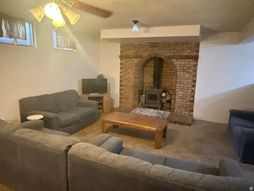 Living space featuring a brick fireplace with a wood-burning stove, two small windows, a ceiling fan with integrated lighting, and carpet flooring