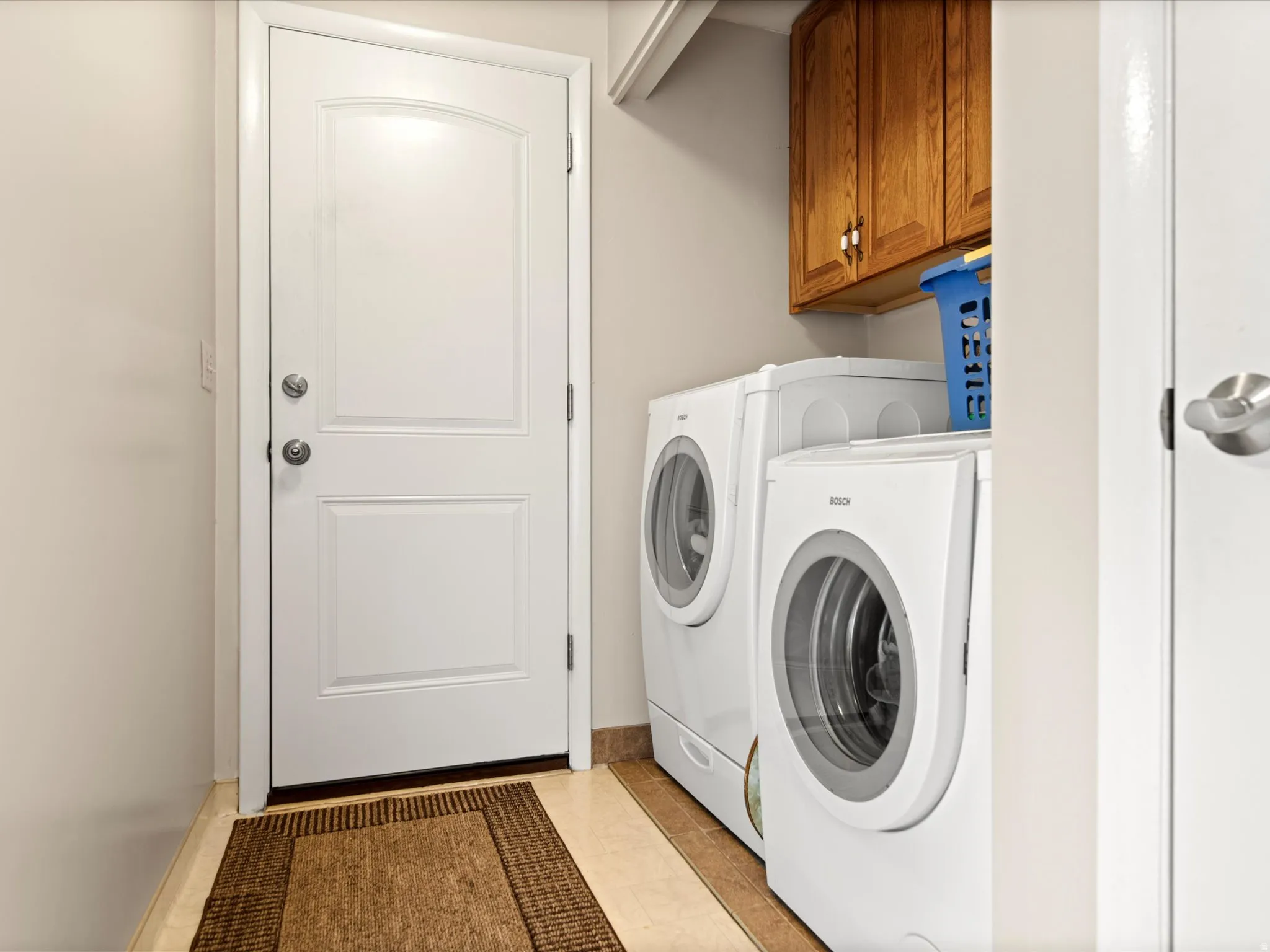 Dedicated laundry area featuring a white paneled door, light-toned wall paint, natural wood-finish cabinetry, and neutral-toned floor tiling