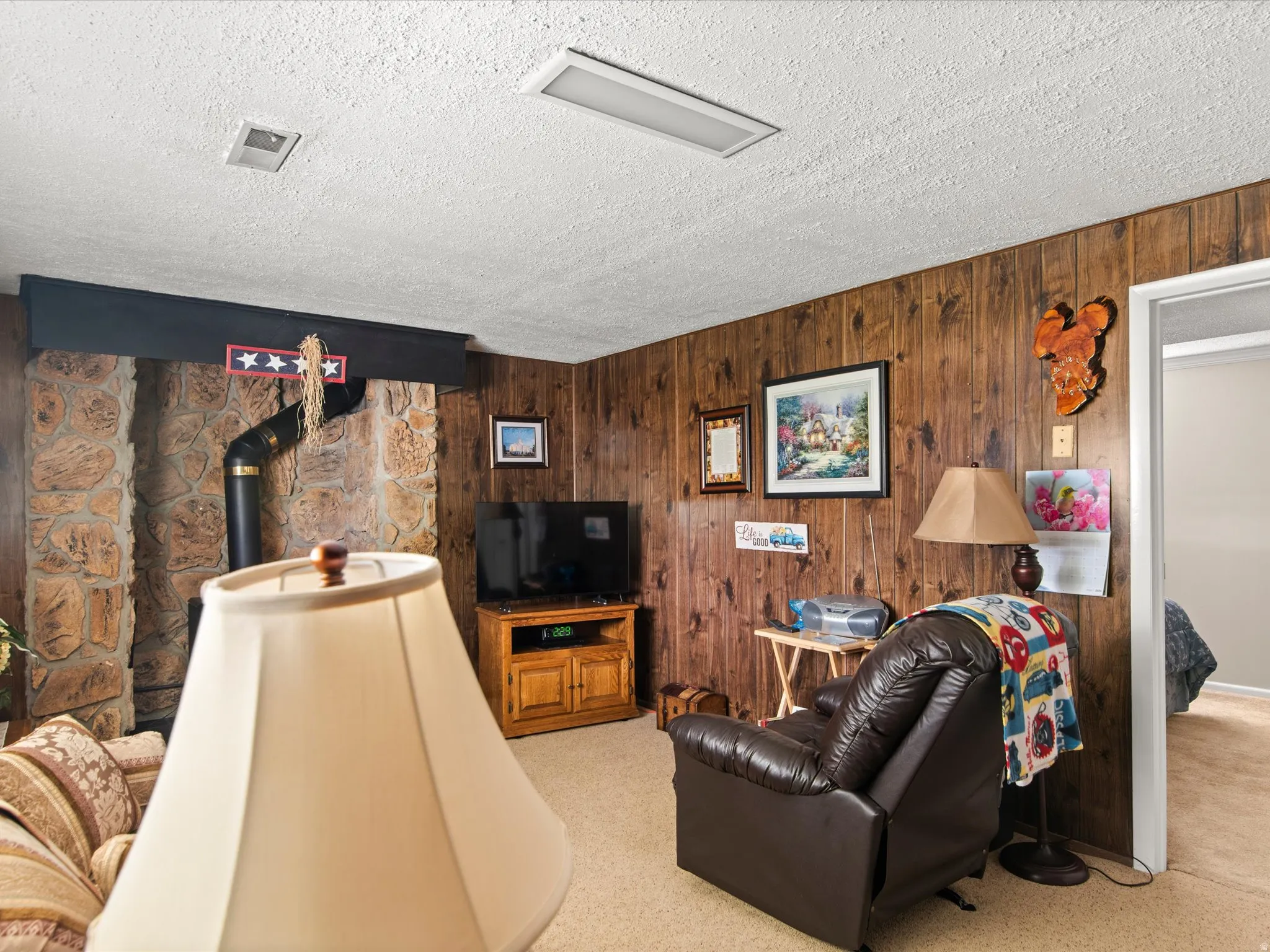Stone-clad fireplace with black stovepipe, wood-finish wall paneling, and neutral carpet flooring