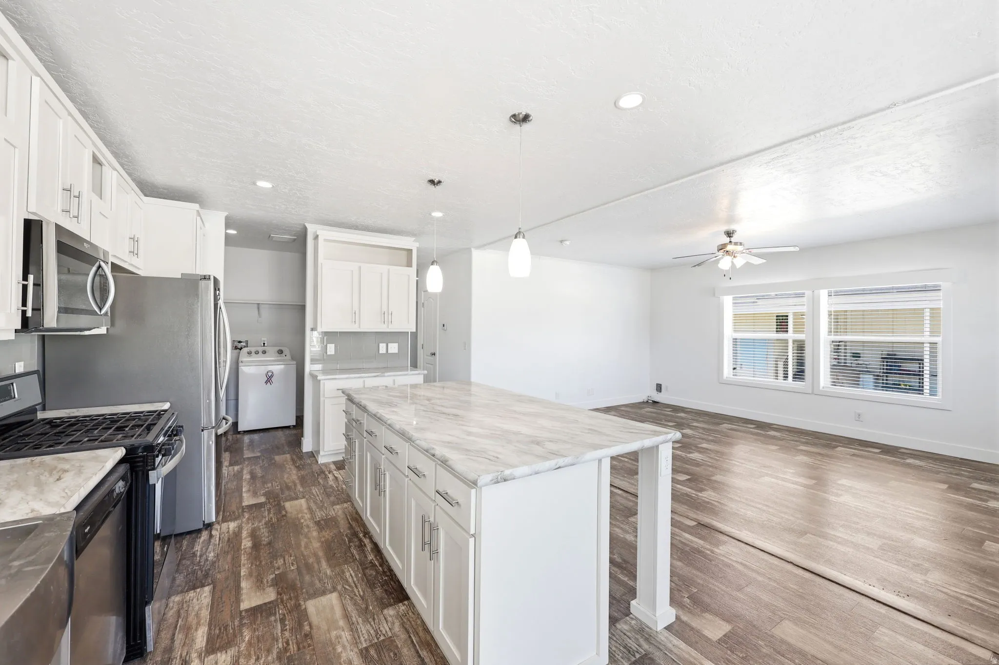 Open-concept living space featuring a kitchen island with a stone-finish countertop, white cabinetry with silver-tone hardware, stainless steel appliances, and wood-finish flooring extending into the living area with a ceiling fan and windows