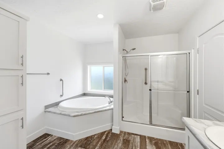 Bathroom featuring a built-in soaking tub with a stone-finish surround, a glass-enclosed shower, wood-finish flooring, and white cabinetry