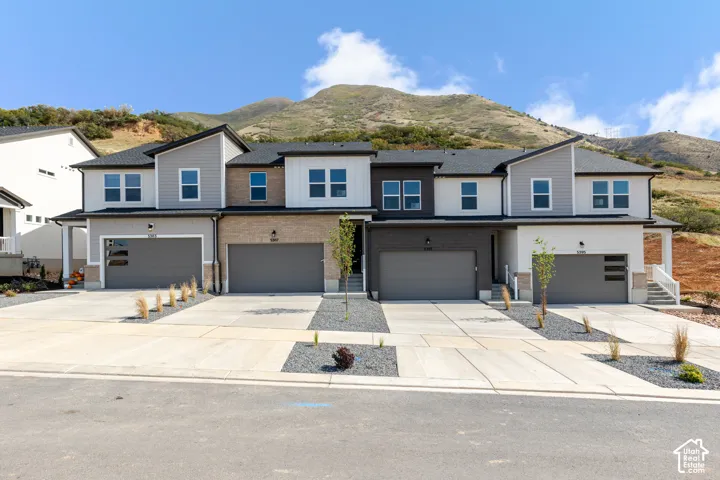 Multi-unit exterior featuring mixed-material siding, stacked-stone accents, shingled roofing, concrete driveways, and mountain-range backdrop