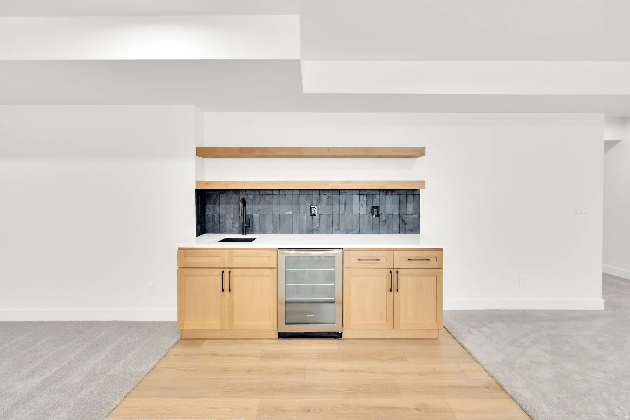 Modern wet bar featuring light wood cabinetry, a white countertop, a black subway tile backsplash, and two floating wood shelves
