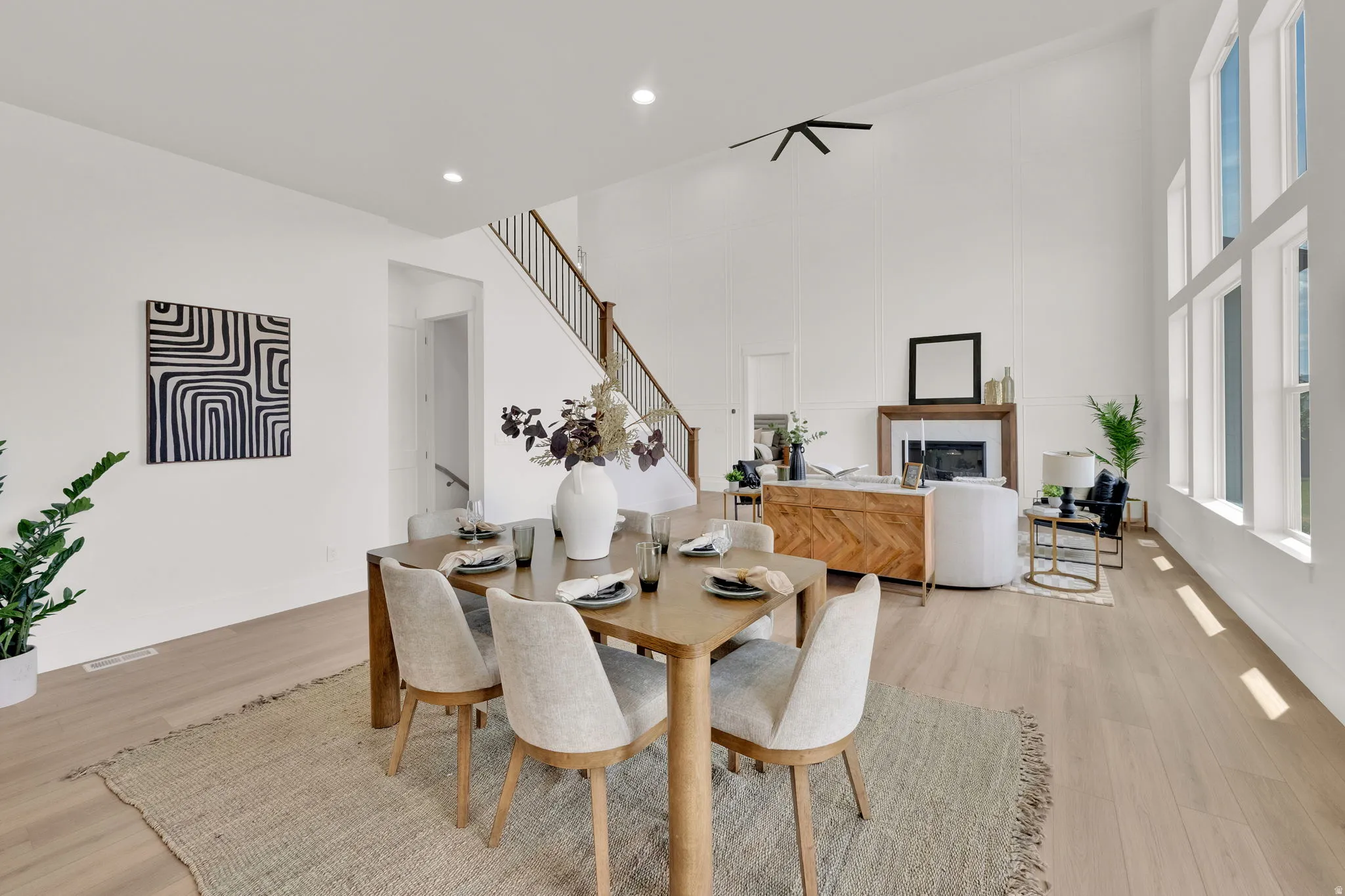 Open-concept living area featuring high ceilings, light wood-finish flooring, a fireplace with a wood mantle, and extensive natural light from multiple large windows