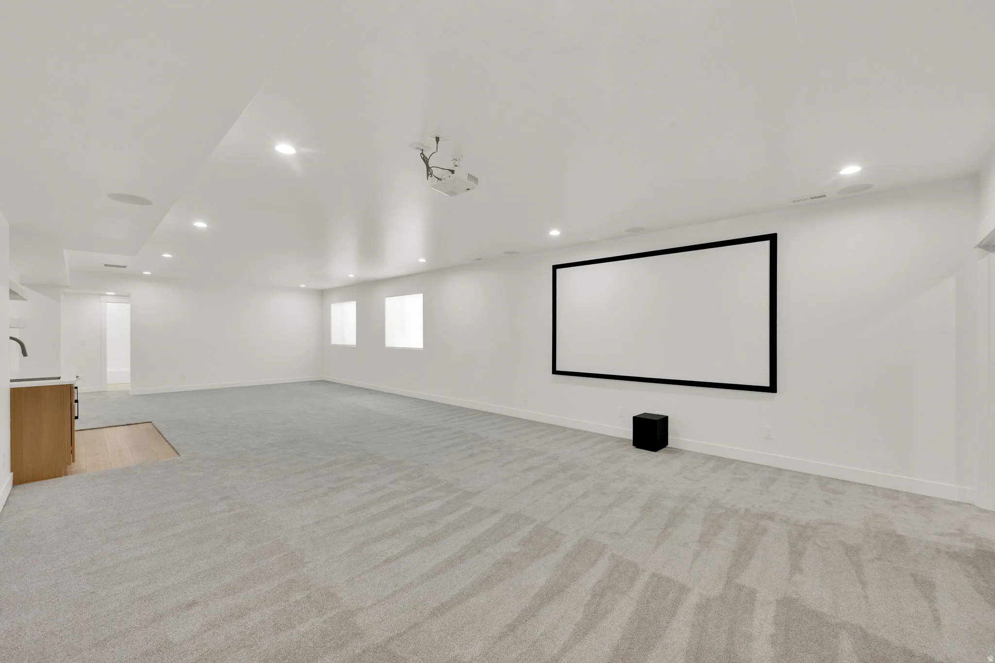 Spacious entertainment room featuring a large projector screen, recessed lighting, light gray carpeting, a built-in wet bar with light wood-finish cabinetry, and white walls