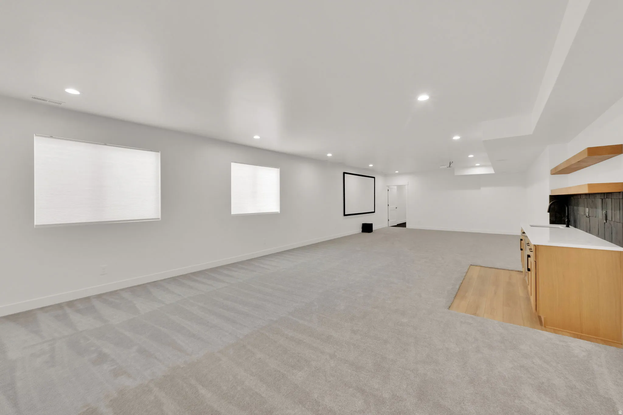Spacious lower-level area featuring light gray carpeting, recessed lighting, and two windows with blinds