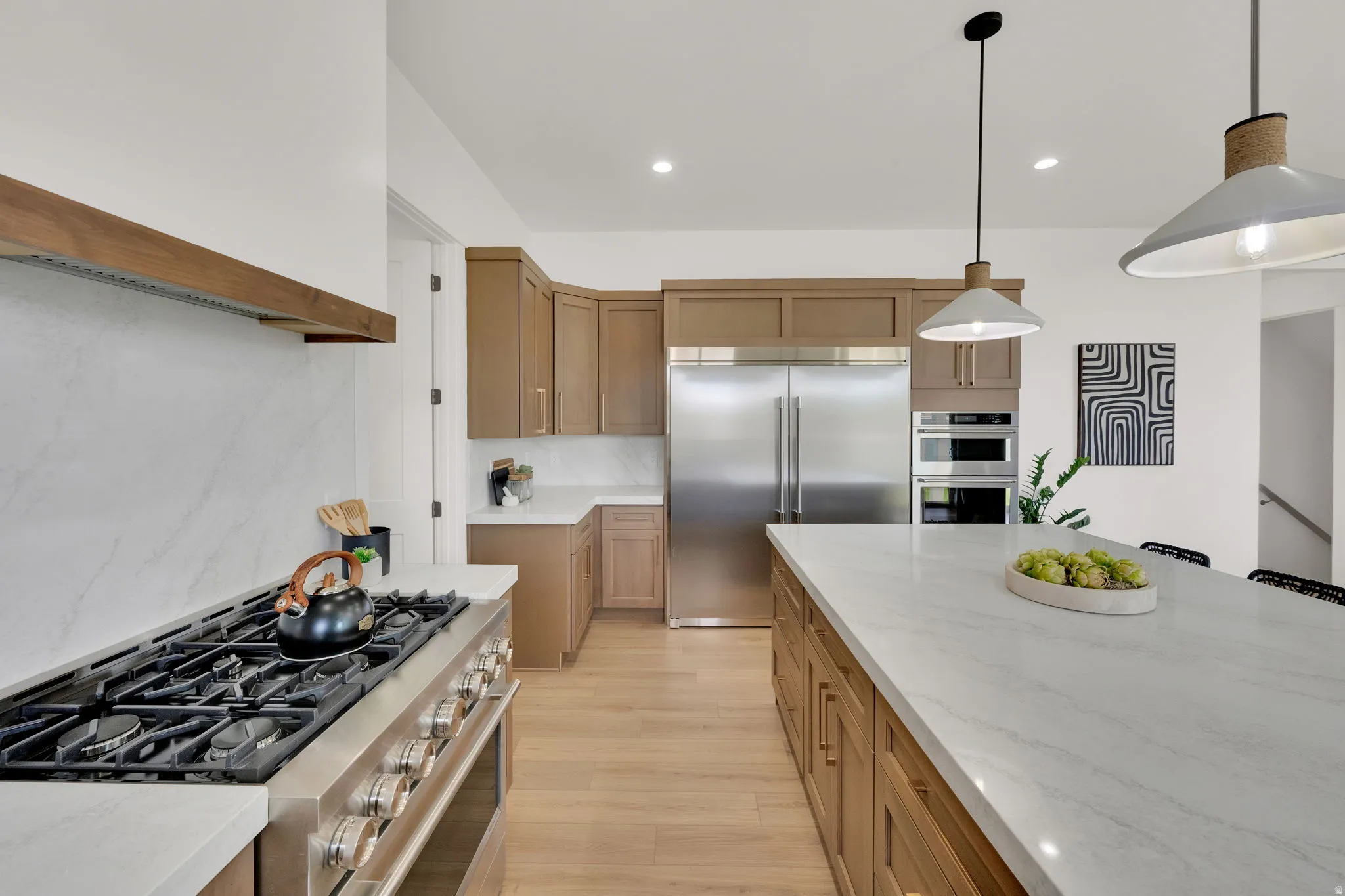 Gourmet kitchen featuring a large island with white stone countertops, stainless steel appliances including a range and double ovens, light wood-finish cabinetry, and wood-finish flooring