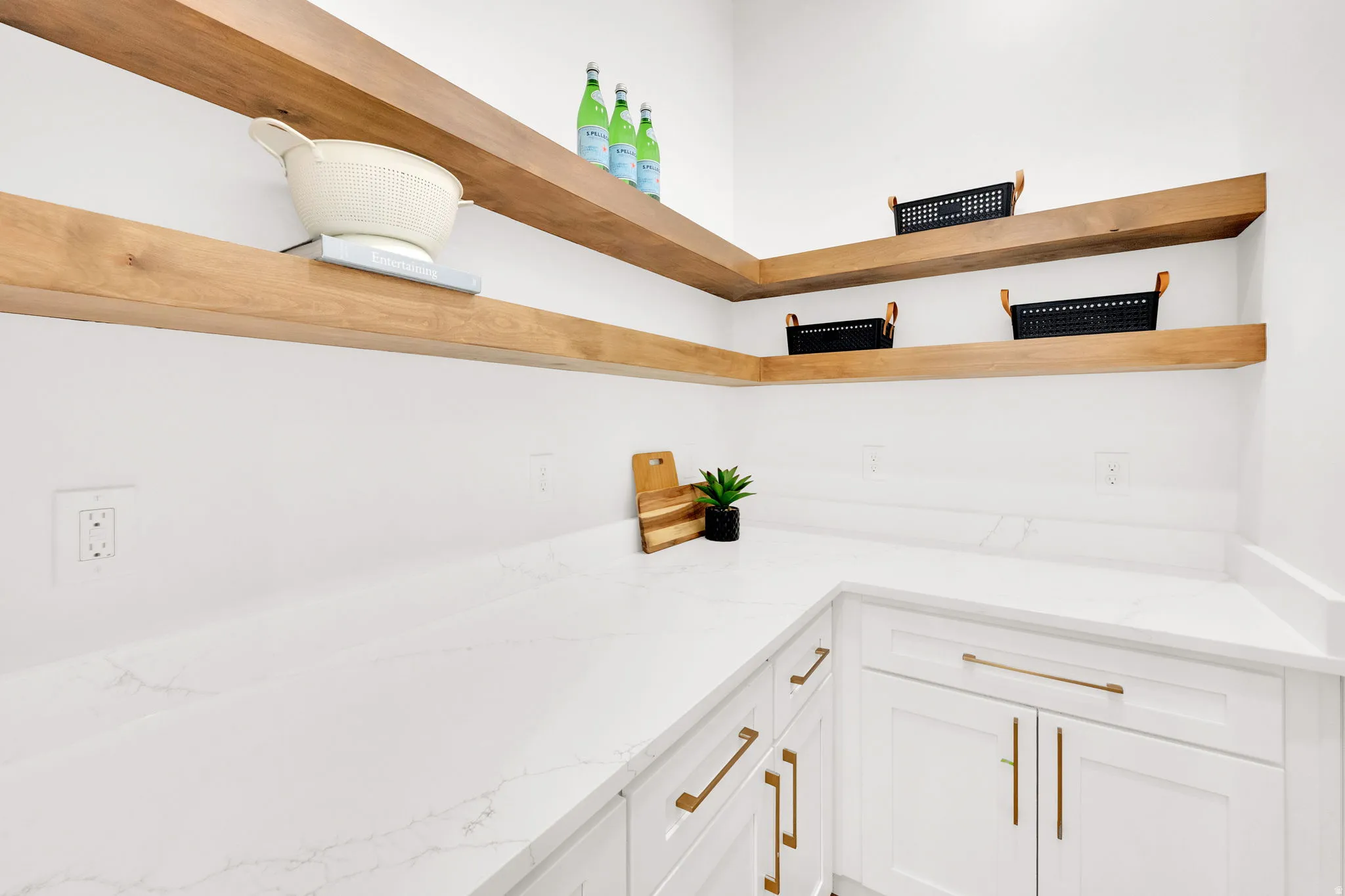 Corner pantry featuring white cabinetry with gold-tone hardware, white countertops, and wood-finish floating shelves