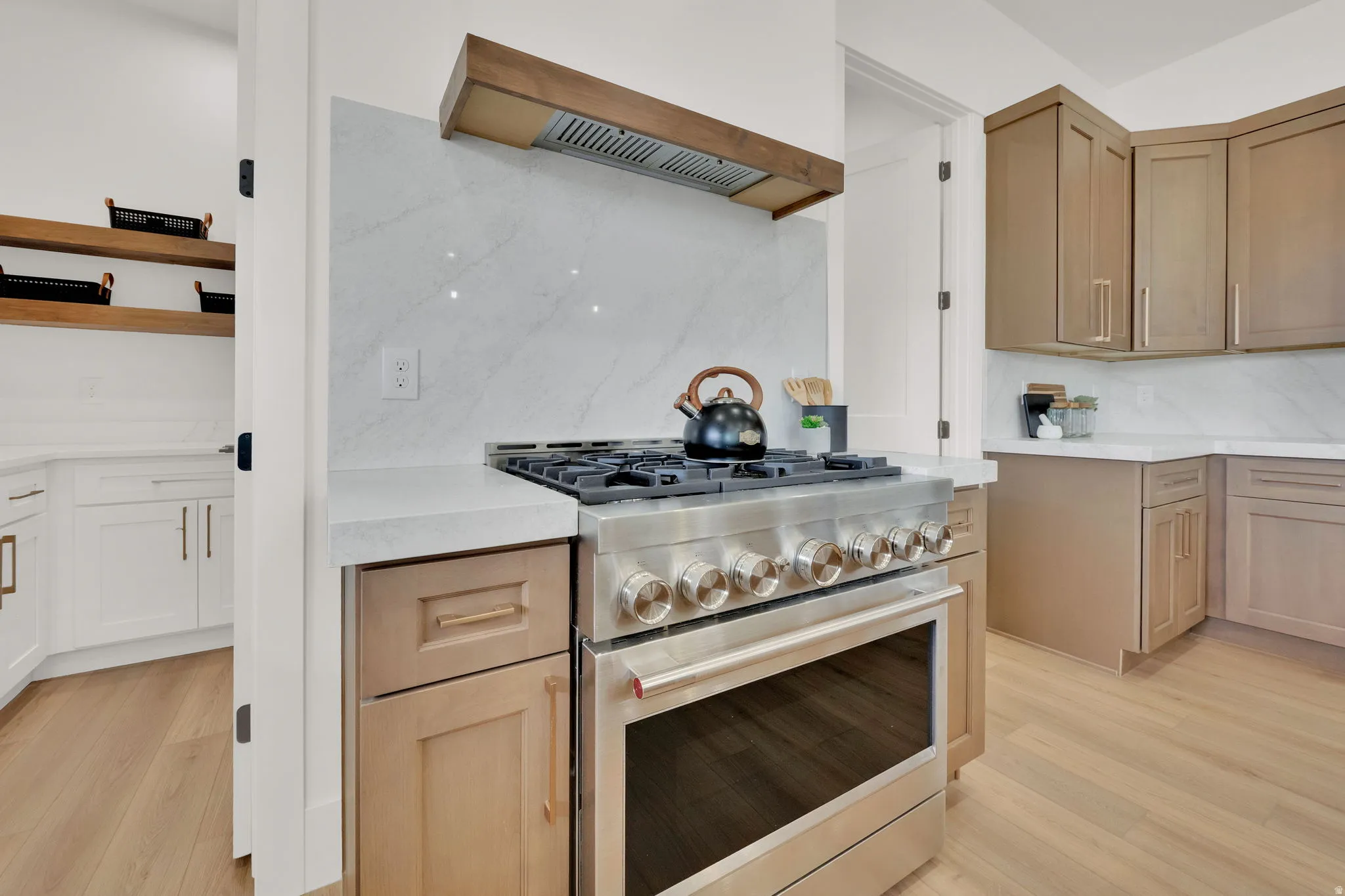 Stainless steel range with a wood-finish vent hood, marble-patterned backsplash, light wood-finish flooring, white countertops, and warm-toned cabinetry