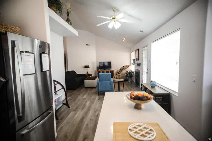 Open-concept living area featuring vaulted ceilings, wood-finish flooring, a ceiling fan, a fireplace with a mantel, and a bright window with blinds