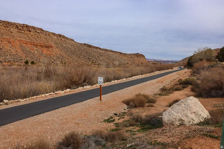 Paved pathway bordered by natural desert landscaping, featuring a speed limit sign, large rock formations, and distant red rock cliffs